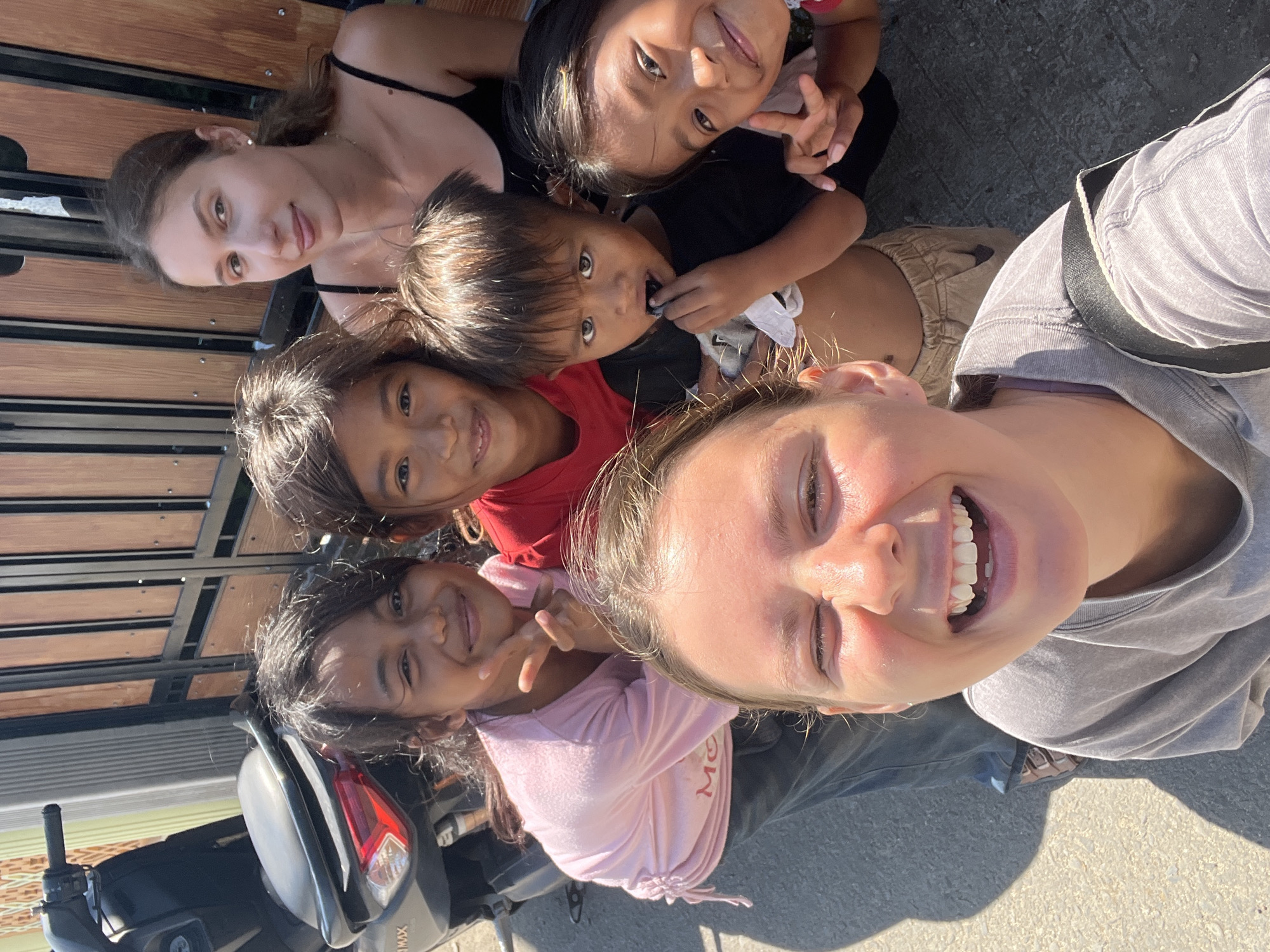In Kertasari, Indonesia, a group of five children and two adults pose for a selfie outdoors. The children, with varying skin tones and dark hair, are arranged in front of a wooden gate. One young girl wears a pink t-shirt, another a red t-shirt, and a young boy is dressed in a black t-shirt. The other two children are partially visible. Two adults are also present, one with light brown hair and a black tank top, the other with blonde hair and a gray t-shirt, with her face filling the foreground of the image. A motorcycle is parked to the left of the group. The ground is paved with asphalt.
