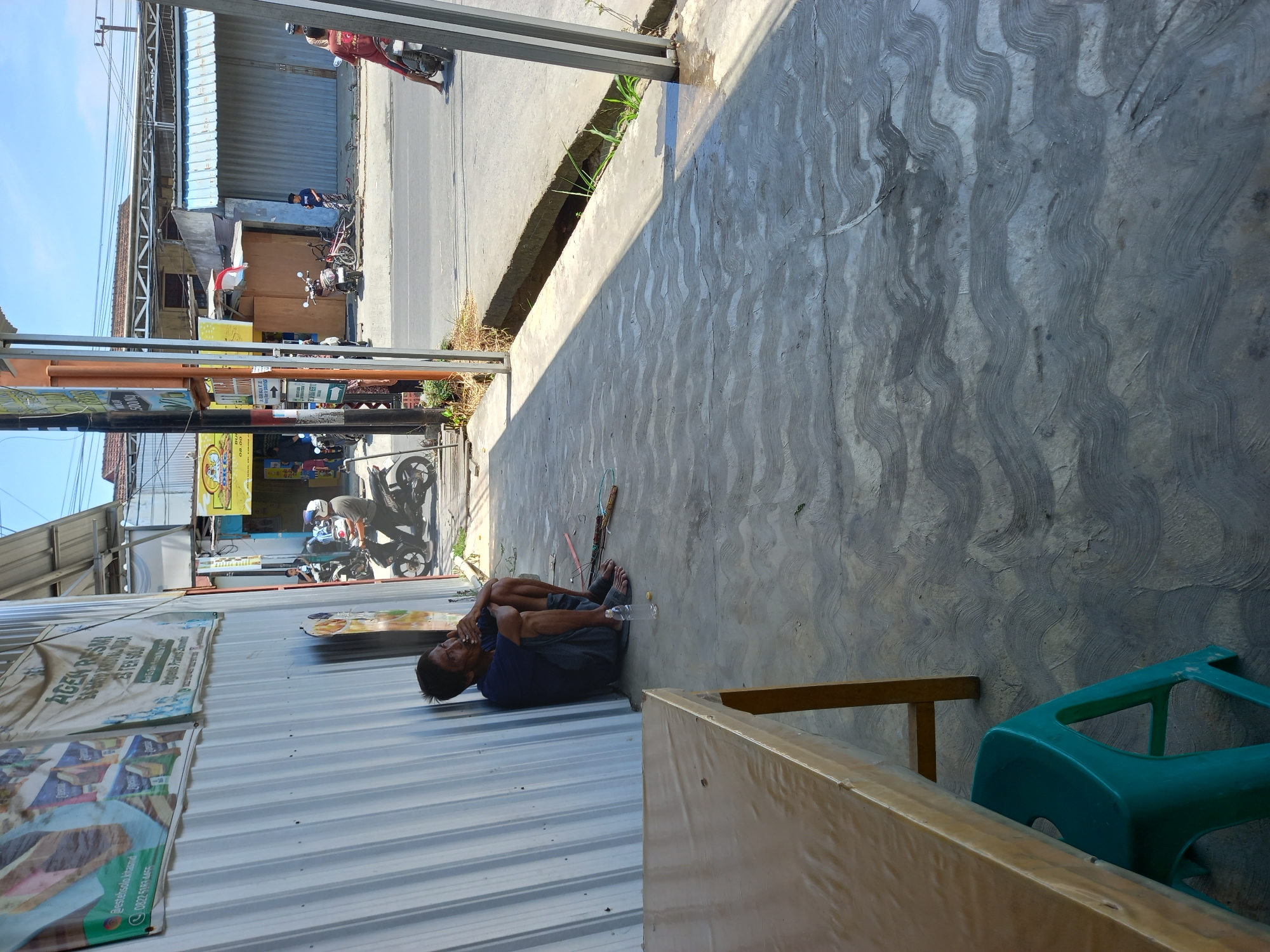 A man is sitting on the ground with his knees pulled up to his chest, leaning against a corrugated metal wall. He is smoking a cigarette and appears to be looking off to his right. He is wearing a dark blue t-shirt and dark shorts, with sandals on his feet. A plastic water bottle is on the ground next to him. In front of him, to the left, is a wooden table covered with a plastic sheet, and a teal plastic stool is underneath the table.
Behind the man, to the right, a street can be seen with several motorbikes and a bicycle. People are present on the street and on the sidewalk. The environment appears to be an outdoor commercial area with buildings and shops lining the street. The ground is paved with concrete that has a wavy, textured pattern. Sunlight casts shadows across the scene. This scene is located on Jalan Pahlawan in Samarinda, Indonesia.