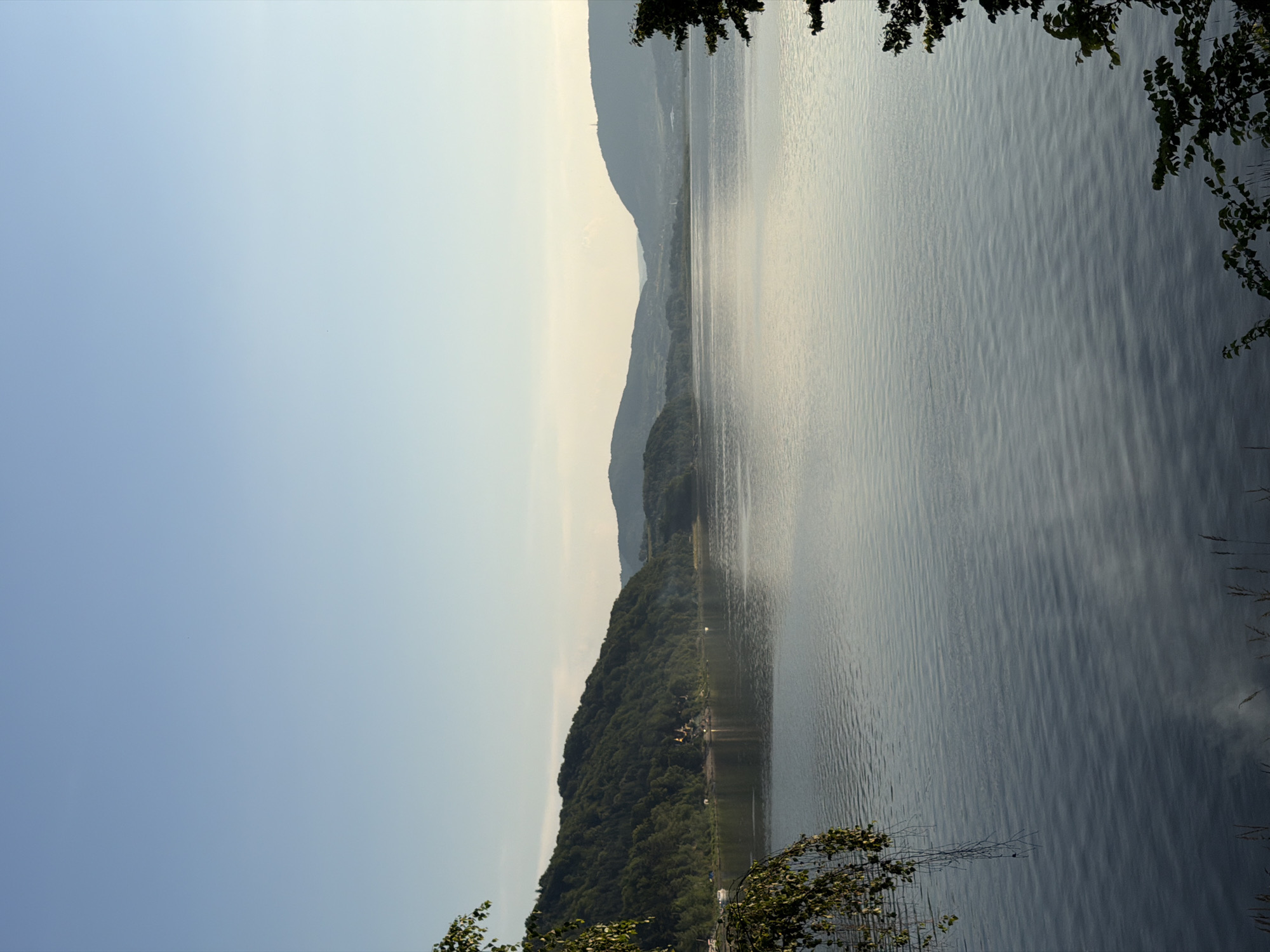 The image depicts a serene lake scene with rolling hills covered in lush green trees in the background. The sky is a clear, pale blue, transitioning to a lighter hue near the horizon, suggesting it is either early morning or late afternoon. The lake's surface is calm, with gentle ripples reflecting the soft light.
In the mid-ground, a densely forested hill slopes down towards the water's edge. A small cluster of buildings, appearing to be houses or a small village, is nestled on the hillside amongst the trees. There are a few boats visible near the shore, indicating potential recreational activity.
Further in the distance, more hills and mountains can be seen, some shrouded in a light haze, giving the scene a sense of depth. The overall impression is one of natural beauty and tranquility, with no visible people or immediate action taking place. The location is identified as the city of Lipie, Poland.