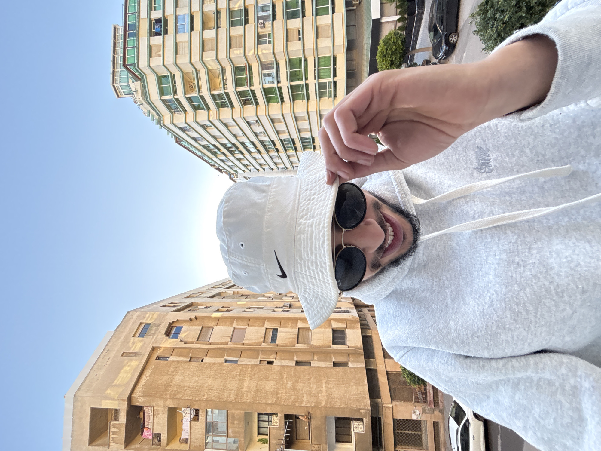 A male individual is captured in a close-up, upward-angled shot against an urban backdrop in Tangier, Morocco. The subject, positioned centrally, wears a white bucket hat with a black Nike swoosh, dark round sunglasses, and a light grey hooded sweatshirt featuring a small embroidered logo and white drawstrings. He has a dark beard and a visible smile, with his right hand adjusting the brim of his hat.
Behind the individual, two multi-story buildings dominate the background under a clear, light blue sky. To the left, a weathered yellowish-brown building with multiple balconies and windows shows signs of age; laundry, including a pink garment, is visible on two balconies. To the right, a more contemporary-looking, cream-colored building features numerous green-framed windows, balconies, and an visible outdoor air conditioning unit.
Below the buildings, a partial view of a street includes two parked vehicles: a white hatchback on the left and a black sedan on the right. Green shrubbery is also present near the street level. The overall scene depicts a daytime urban environment.