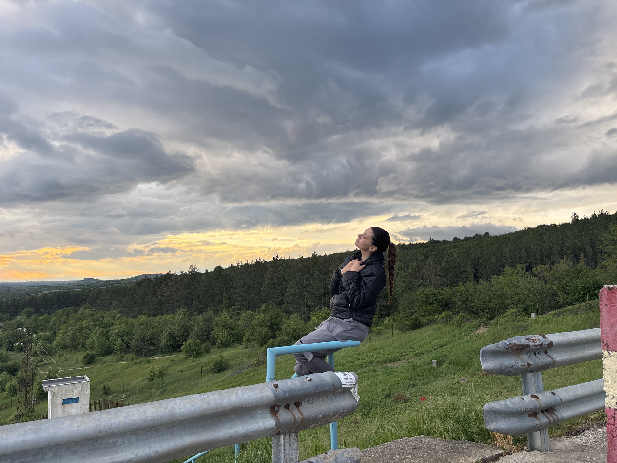 A woman is seated on a blue metal railing on an overlook in Ognyanovo, Bulgaria. She is wearing a black jacket and grey pants, with her eyes closed and head tilted back towards the sky, which is filled with dark, cloudy formations. In the background, there is a forested landscape with rolling hills. In the left lower portion is a small, concrete, windowed structure near electrical lines, and a metal guardrail with paint is present.