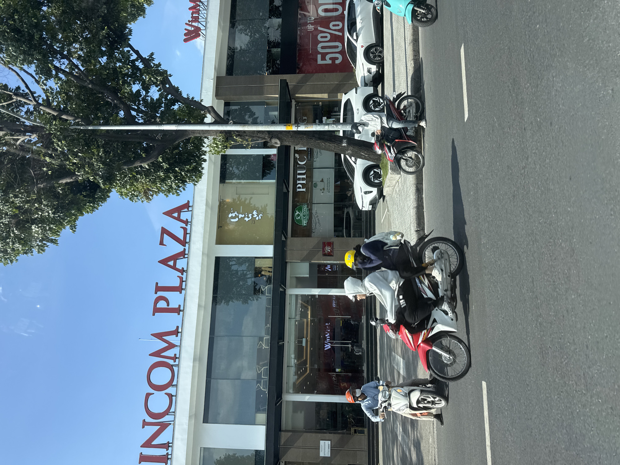 This image depicts a street scene in Thuận An, Vietnam, with a prominent "VINCOM PLAZA" sign visible on the facade of a modern building. The building features large glass windows and a dark awning above the entrance. Several people are on motorcycles, navigating the street and sidewalk. One motorcycle in the foreground carries two individuals, one wearing a white hoodie and the other a dark hoodie with a yellow helmet. Another person on a motorcycle to the left is wearing a blue shirt and an orange helmet. Parked cars, including white sedans and SUVs, are visible on the side of the road, some with visible branding like "WinMart." A large tree with dense foliage stands near the building, and a utility pole is also present. The sky is clear and blue. The overall scene suggests a bustling urban environment on a sunny day.