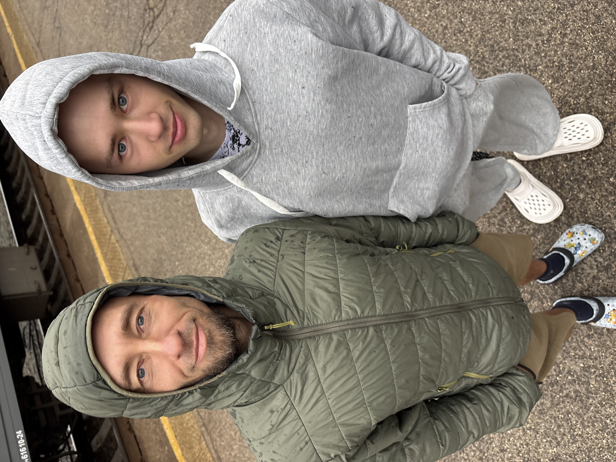Two men in hooded jackets stand closely together on a textured ground with a yellow safety strip at the edge. The man in the gray hoodie is at the top of the frame, wearing white slip-on shoes. The man in the green quilted jacket is below him, wearing light brown pants and black socks with patterned slip-on shoes. The background contains elements suggesting an urban environment, potentially a train platform, indicated by a dark object with visible numeric text. The location is identified as Saratov, Russia.