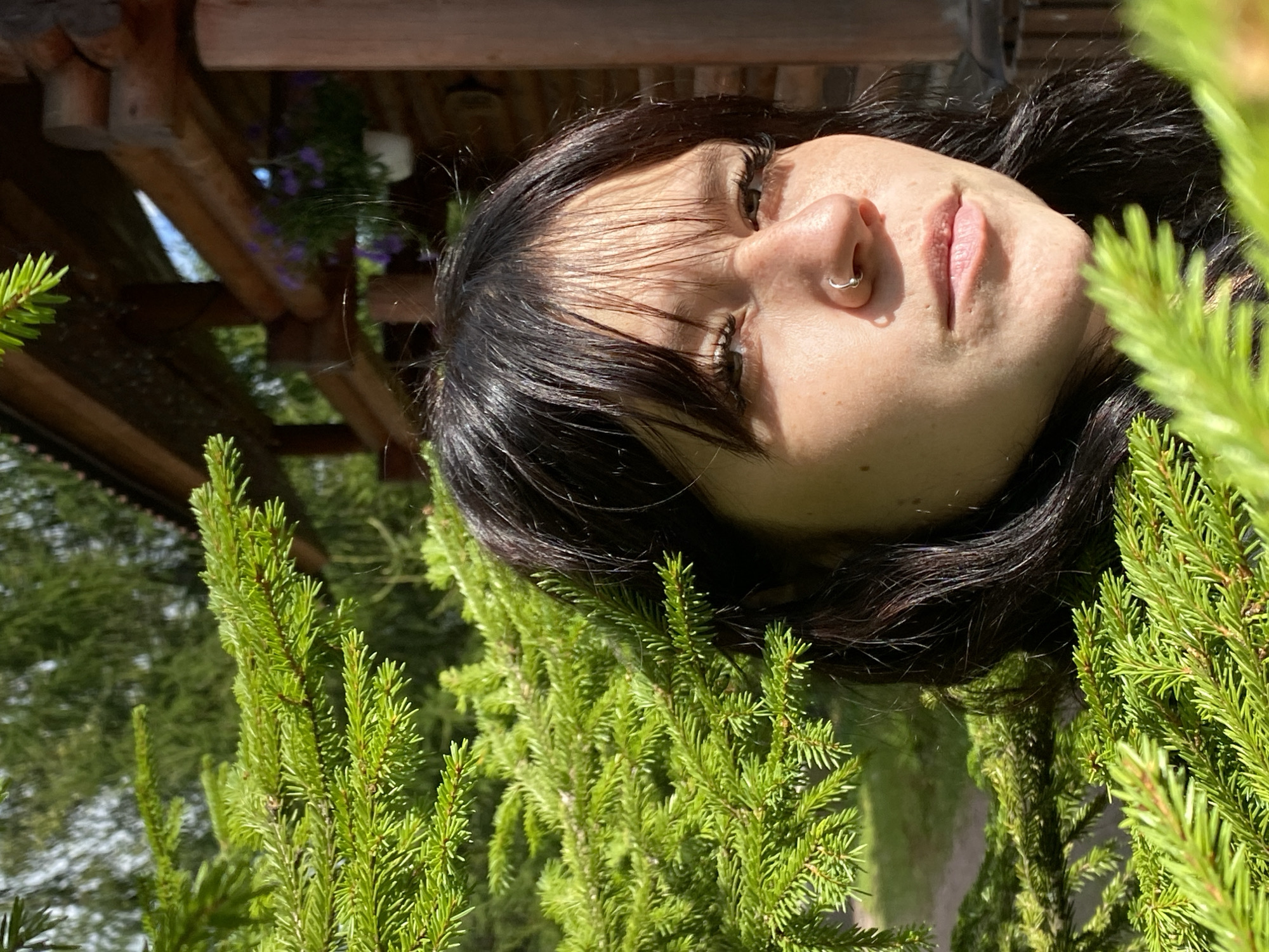 A young woman with dark hair and freckles is partially obscured by a spruce tree in front of a wooden log cabin. She is wearing a nose ring. The cabin has a porch with hanging flower baskets. The scene is outdoors and appears to be in a rural or wooded area. It is daytime, with sunlight filtering through the trees. The location is Krasnoflotskoe, Russia.