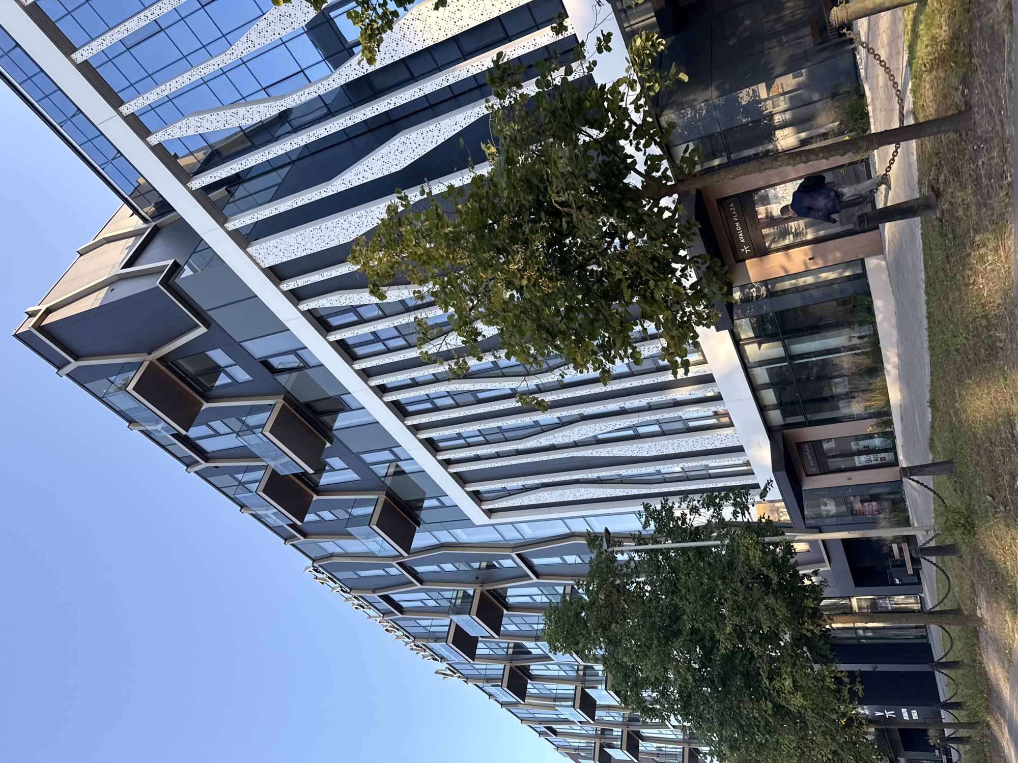 The image shows the modern Avalon Plaza building in Tallinn, Estonia. The building features a distinctive facade with a mix of dark gray panels, white patterned vertical accents, and numerous glass windows and balconies. Tall green trees line the sidewalk in front of the building, casting shadows. A person in a dark jacket and light-colored pants is walking away from the entrance of the building, which has a sign reading "AVALON PLAZA". A chain fence with bollards separates the sidewalk from a grassy area. The sky is clear and blue.