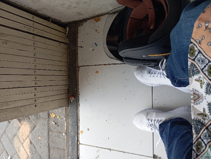 The scene is a downward-facing, first-person perspective at a doorway or entrance in Legok, Indonesia. A person is seated or standing, with their lower legs and feet visible. They are wearing blue denim jeans and white lace-up athletic shoes. A portion of patterned fabric, featuring geometric and floral designs in shades of brown, green, and white, is visible in the immediate foreground. To the right of the person's right foot, a black motorcycle helmet with a brown interior lining and a yellow stripe is resting on the floor. The flooring consists of light-colored, possibly white, ceramic tiles that show some dirt and scattered debris, including small paper fragments, a blue and white plastic cap, and orange crumbs. A dark metal threshold separates the tiled area from an exterior paved surface composed of interlocking grey blocks. Partially open, a light-colored, accordion-style metal security grille or gate, showing signs of wear, is positioned along the threshold, extending across the upper portion of the frame. The outdoor pavement area also contains scattered debris, including cigarette butts, yellow-orange paper fragments, and a small red and white object near the threshold.