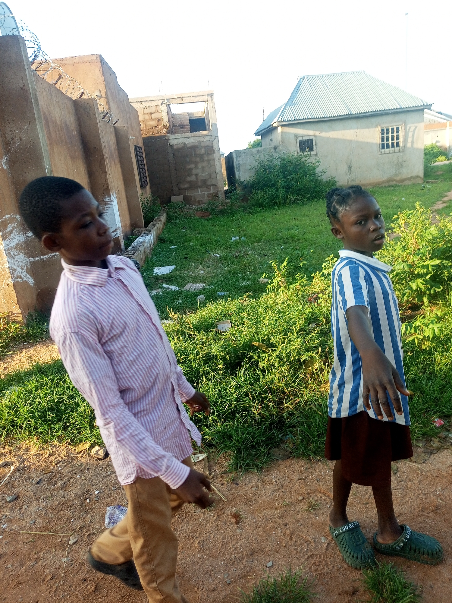 In Tempure, Nigeria, two children, a boy and a girl, walk on a dirt path beside a grassy area. The boy, on the left, wears a pink and white striped long-sleeved shirt and tan pants. He is looking away from the camera, to his left. The girl, on the right, wears a blue and white striped short-sleeved shirt and a dark skirt. She is looking at the camera and has her right hand extended forward. Behind the children are several buildings, including a partially constructed structure with brickwork and a finished building with a corrugated metal roof. The environment appears to be rural or on the outskirts of a settlement, with scattered vegetation and unpaved ground. The lighting suggests it is daytime, likely late afternoon or early morning, given the long shadows.