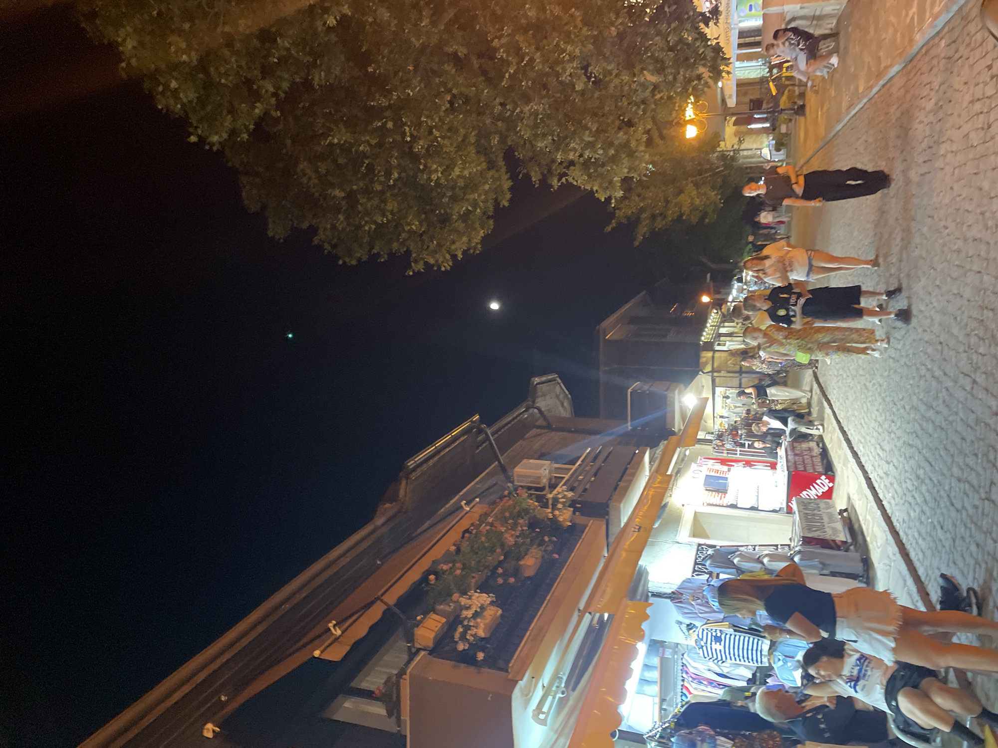 This is a nighttime photograph of a street in Nesebar, Bulgaria, likely the old town, characterized by cobblestone pavement and traditional architecture. Several shops line the street, displaying clothing and jewelry for sale, illuminated by awning lights and street lamps. A group of people are walking along the street, some browsing the shops, while others are seated. The sky is dark with a visible moon and a faint green light, possibly an airplane or satellite. The overall scene depicts a lively evening atmosphere with tourists and locals enjoying the streetscape and shops.