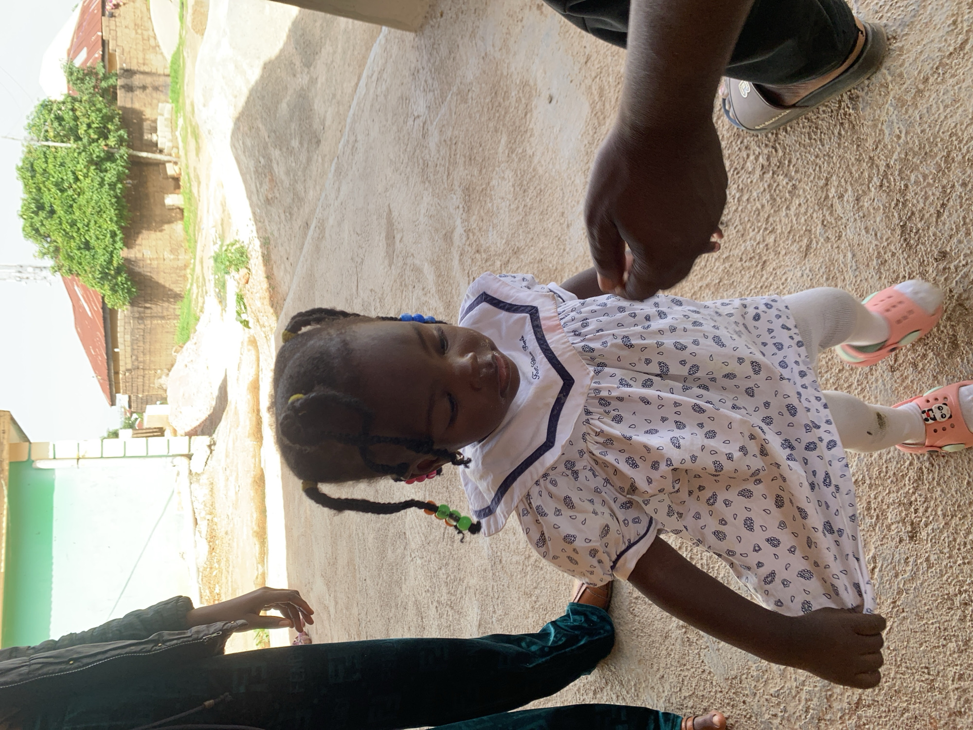 A low-angle, close-up shot shows a young Black girl in a white dress adorned with a blue and white pattern. She has dark hair braided into pigtails, decorated with colorful beads. She is wearing white stockings and pink sandals. The girl is looking down, with her eyes closed, and appears to be sleepy or relaxed.
Her right hand is holding onto the hem of her dress, and her left arm is slightly bent. Her legs are visible, with her right foot wearing a pink sandal. Nearby, a person's dark-skinned hand is visible, holding onto the girl's arm. Another person's legs and feet are visible at the bottom of the frame, one wearing dark green trousers and the other in a brown sandal. A person's hand is also seen near the girl's elbow, wearing a brown sandal.
The ground is a sandy, textured surface, possibly a courtyard or outdoor area. In the background, there are glimpses of a building with a reddish-brown corrugated metal roof and a wall made of rough-hewn stones or bricks. A lush green tree with dense foliage is also visible. The lighting suggests it is daytime, with shadows cast on the ground, indicating sunlight. The overall impression is of a casual, everyday moment, possibly in a residential area in Jos, Nigeria. No visible text is present in the image.
