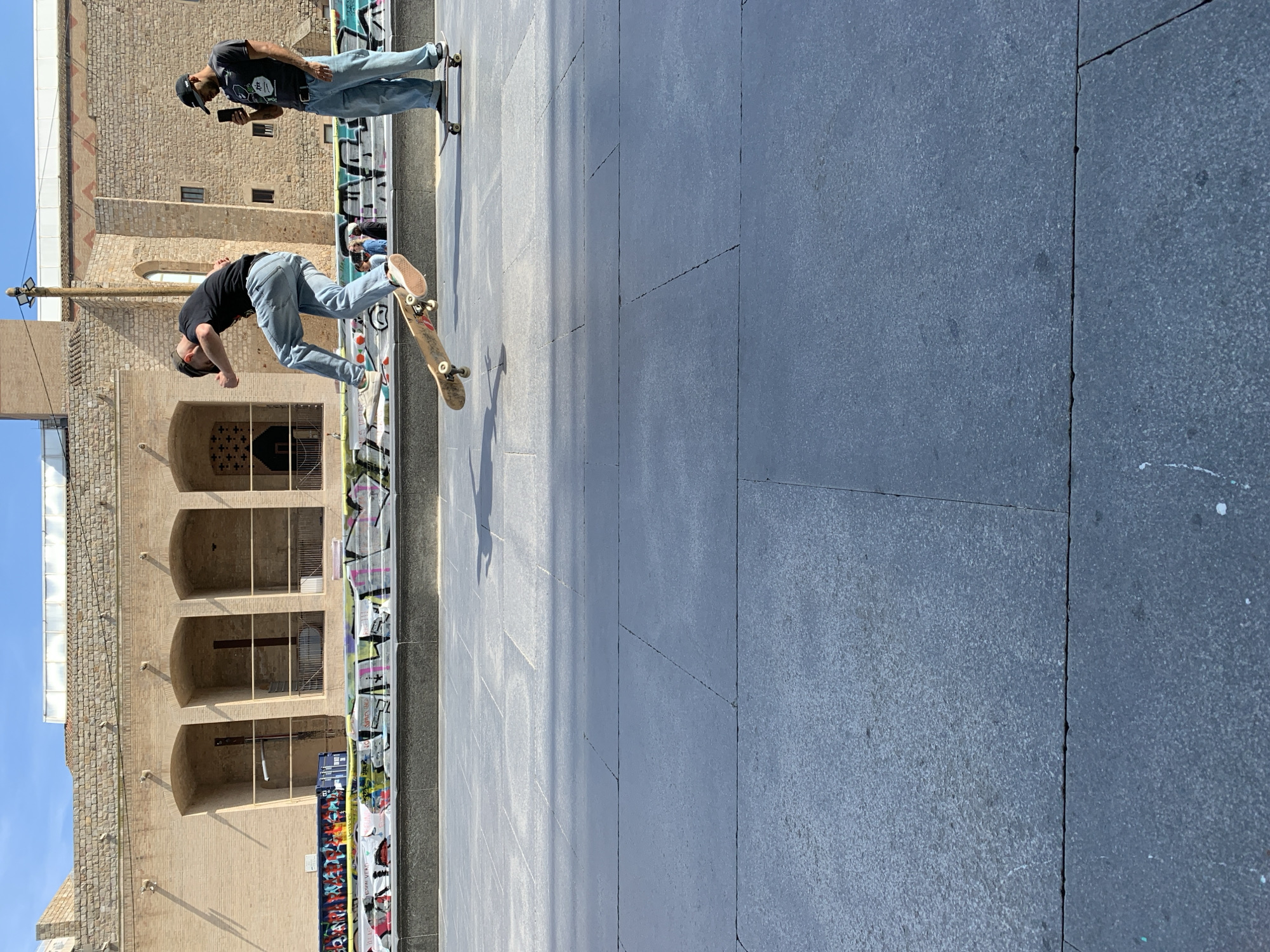 The image shows two skateboarders in Barcelona, Spain. One skateboarder is in the air, performing a trick on his board, while the other is standing on his board looking at his phone. The building behind them is an old, beige stone structure with arches. Graffiti lines the building's base, and the plaza itself is paved with large, gray stones. The environment appears to be sunny.