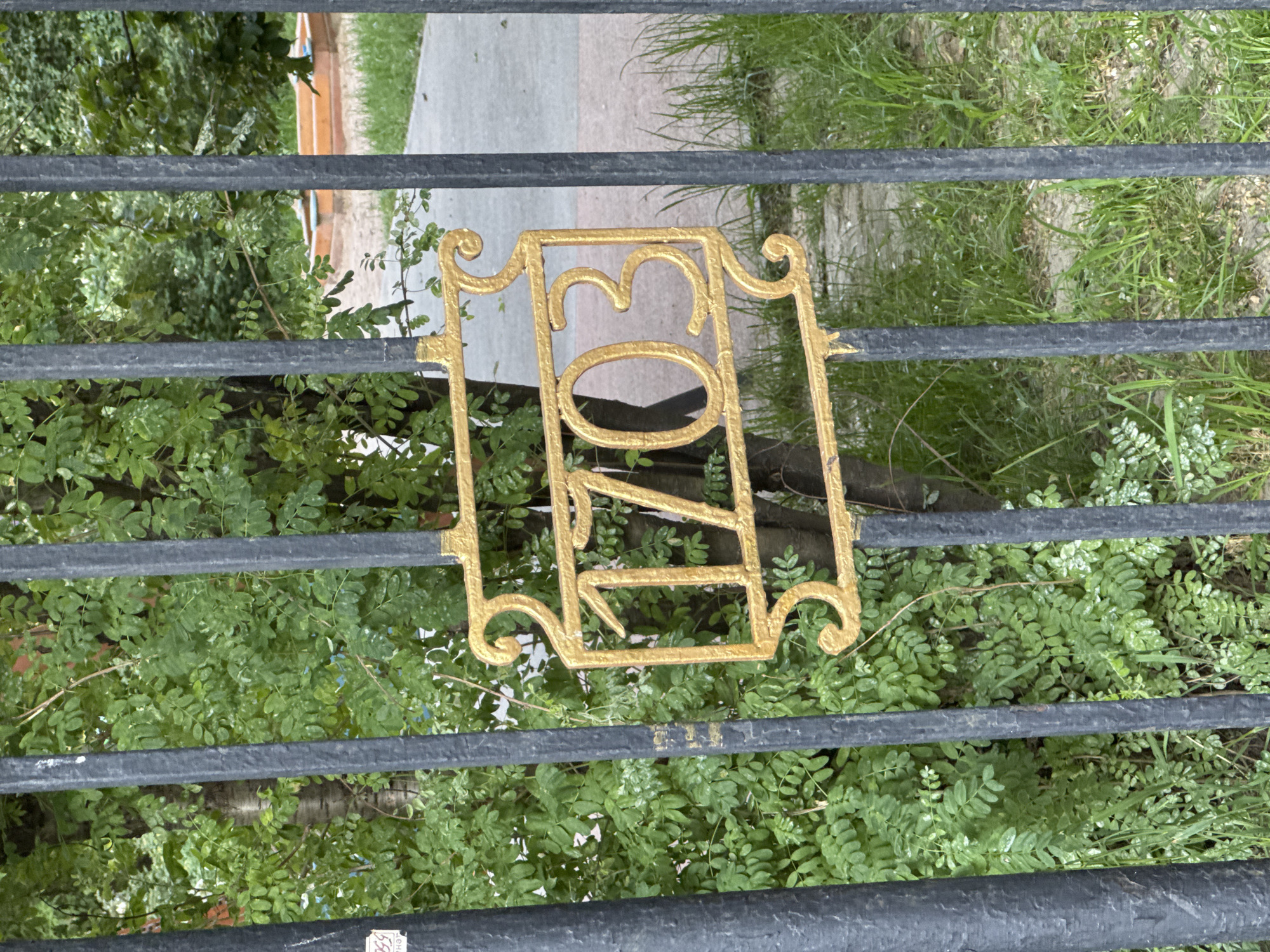 A gold-colored metal plaque with ornate scrollwork and the number "1703" is attached to a black metal fence. The fence is vertical and appears to be made of wrought iron. Behind the fence, there is lush green foliage, likely trees and bushes. In the background, a concrete path is visible, with some patches of dry grass. The lighting suggests it is daytime. The image is taken from a slightly low angle, looking up at the plaque and fence. The plaque's design and the number "1703" may indicate a historical building or a specific address in Saint Petersburg, Russia, as the year 1703 marks the founding of the city.