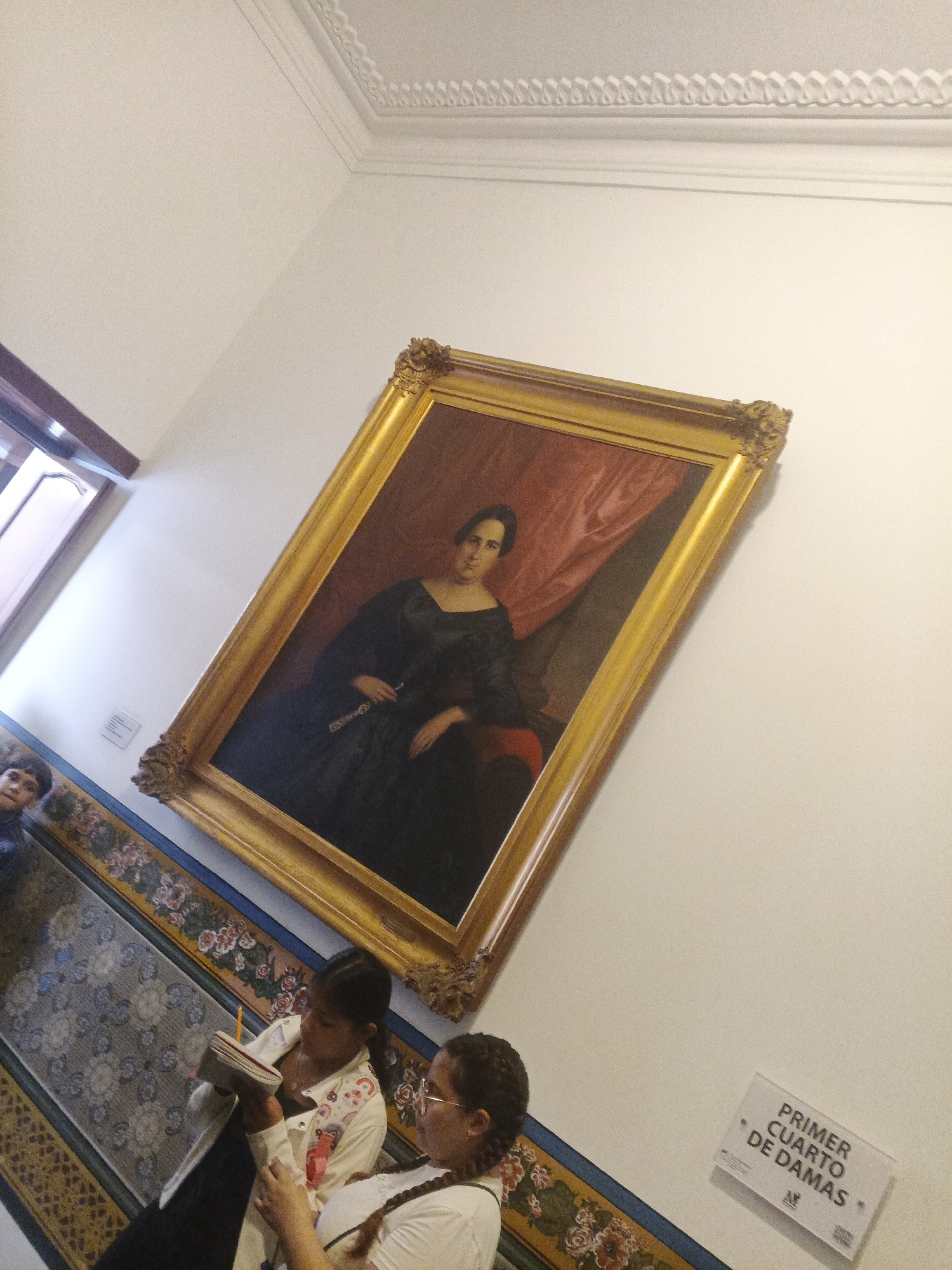 The image shows a room in an unknown location. On the wall, there is a large portrait in a gold frame of a woman in a black dress with a red drape behind her. Below the portrait, there is a tile border with floral patterns. Two people, possibly an adult and a child, stand in front of the tile border. The child is writing in a notebook, and the adult is next to them. A sign reading "Primer Cuarto de Damas" is visible on the wall to the right of the tile border. The room has a white ceiling with decorative molding.