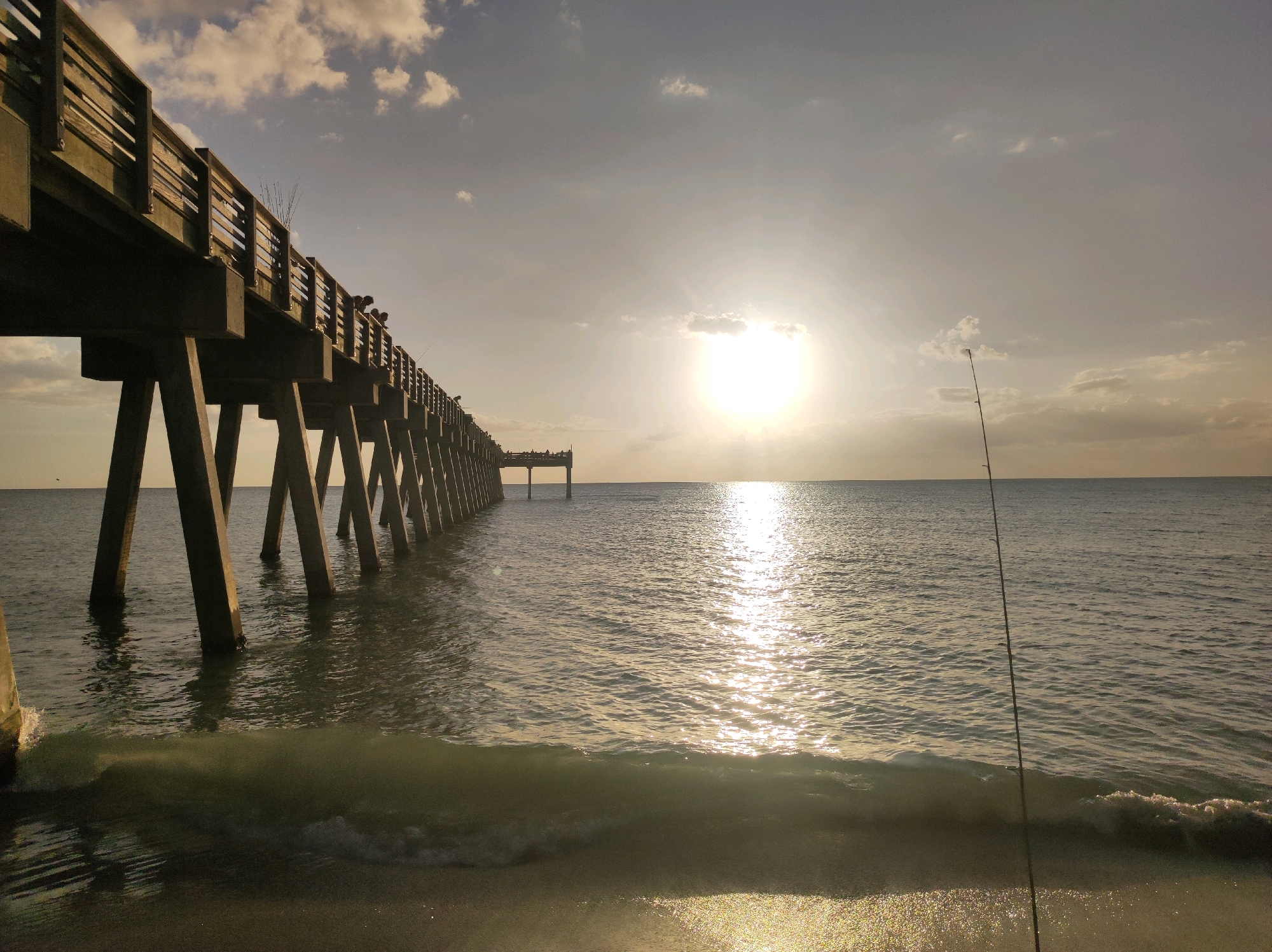 The image captures a serene sunset over the ocean, viewed from the Venice Fishing Pier in Venice, Florida, United States of America. The pier, constructed with weathered wooden planks and sturdy concrete pilings, stretches diagonally across the frame, extending into the tranquil water. Several figures, presumed to be fishermen, are visible along the pier, engaged in their activity as indicated by the presence of fishing rods. A single fishing rod stands upright on the sandy shore in the foreground, its line disappearing into the ocean. The sun, a brilliant orb, hangs low in the sky, casting a shimmering golden reflection across the gently lapping waves. Wispy clouds dot the pale, warm-toned sky, enhancing the peaceful ambiance of the scene. The water exhibits a calm demeanor, with small waves breaking softly on the shore. The overall impression is one of tranquility and natural beauty, characteristic of a coastal sunset.