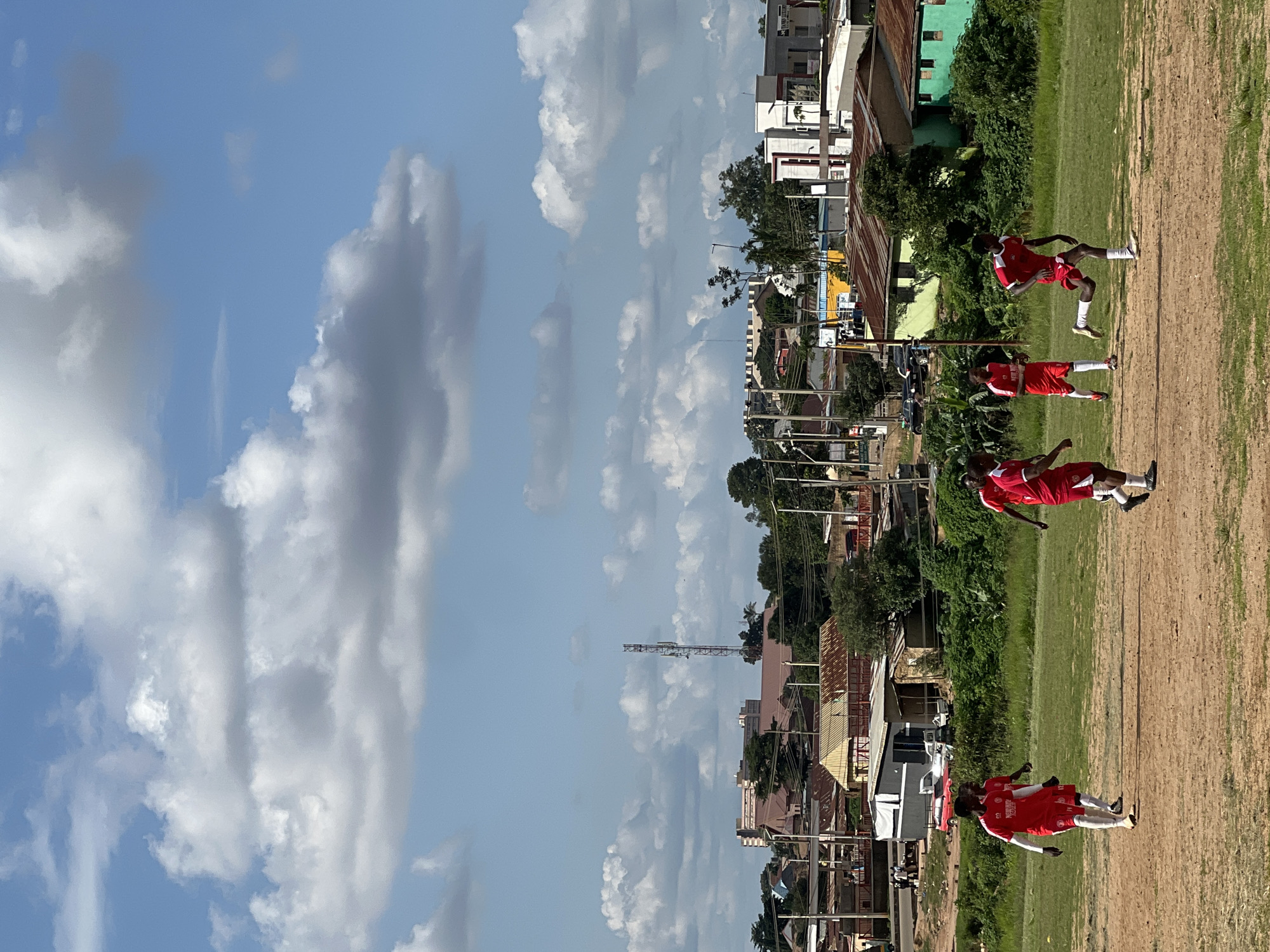 A group of young men in red soccer uniforms are playing soccer on a dirt field in a neighborhood of Jos, Nigeria. The sky is blue with scattered white clouds. In the background, there are buildings, trees, and a telecommunications tower. The players are running and kicking the ball. The scene is illuminated by daylight.