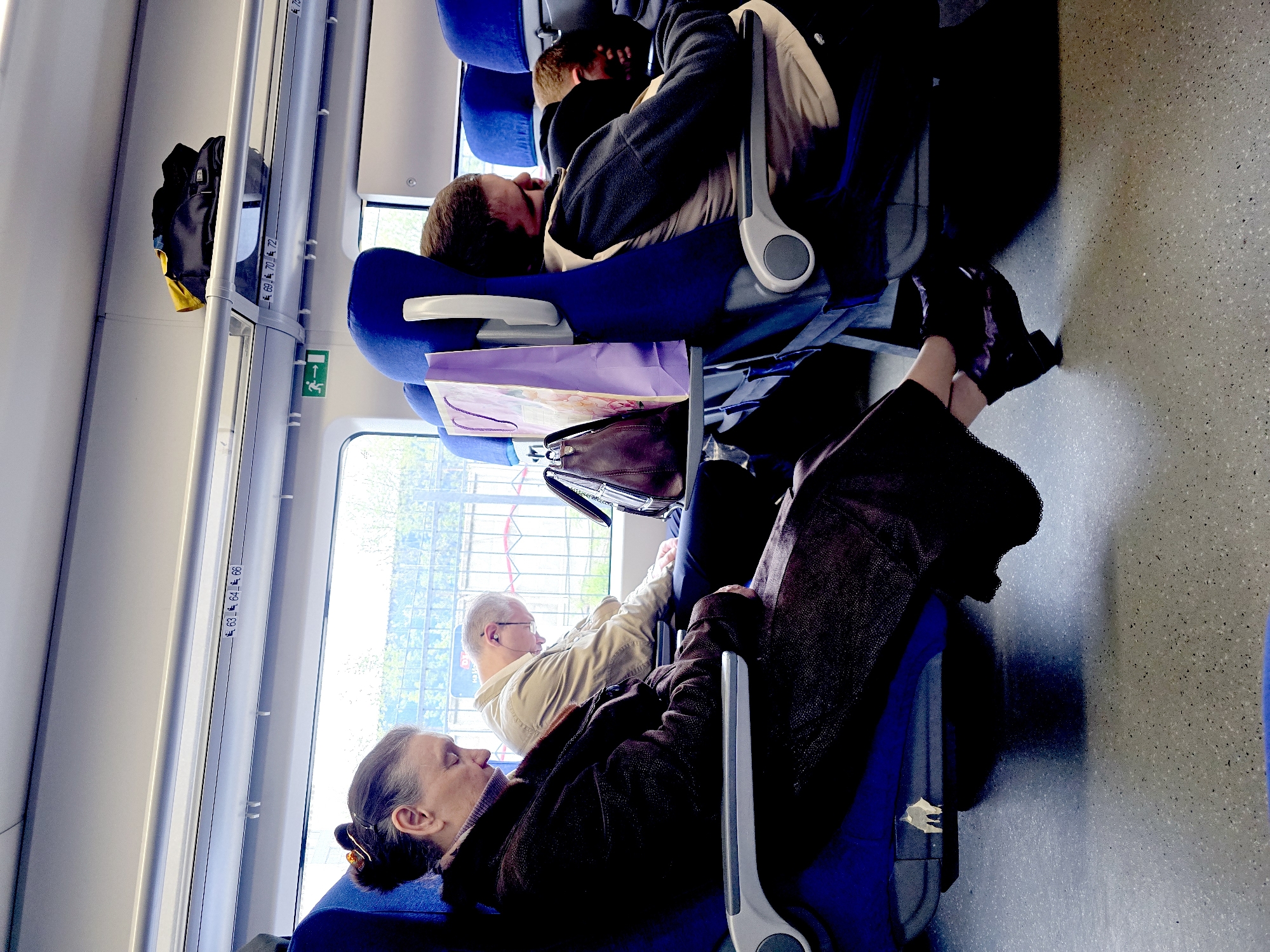 The image depicts the interior of a train carriage. Several passengers are visible, some appearing to be asleep. The seating is blue, and overhead luggage racks are present. A bag is stowed on one of these racks. The flooring has a speckled gray pattern. Outside the window, hints of greenery suggest an urban or suburban environment. The scene appears to be in Unknown, Unknown.