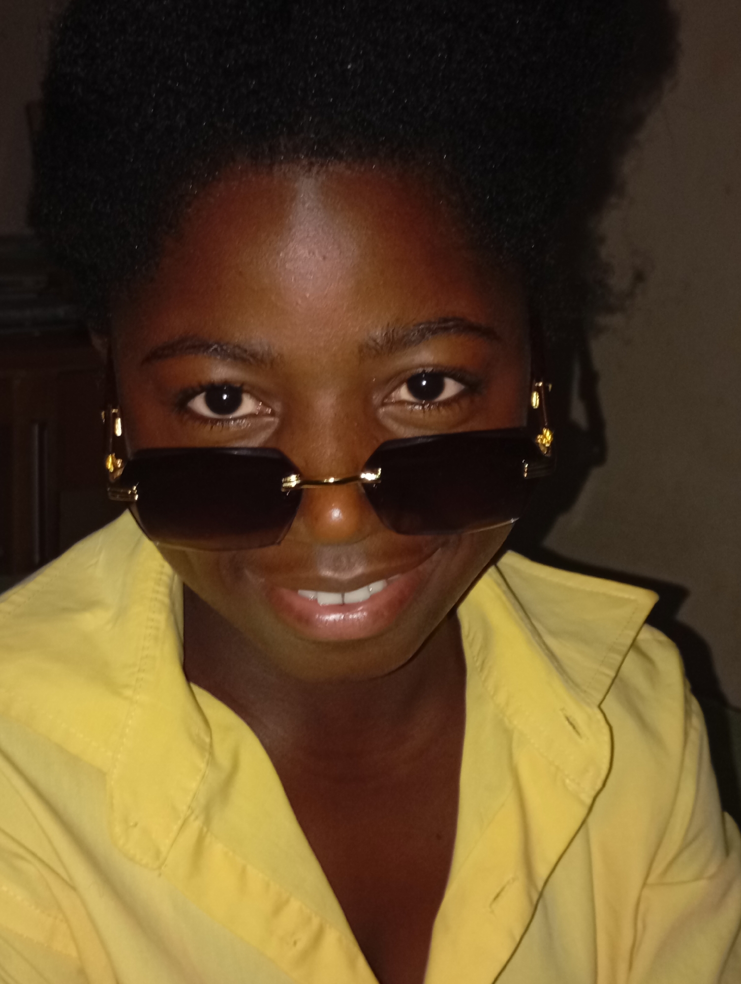 A close-up, medium-lit indoor shot shows a dark-skinned person wearing dark sunglasses with a gold frame and a bright yellow button-down shirt. The person appears to be in front of a dark background. While it is impossible to determine the exact location in Jos, Nigeria, the image provides no specific environmental cues to place the subject within a particular setting.