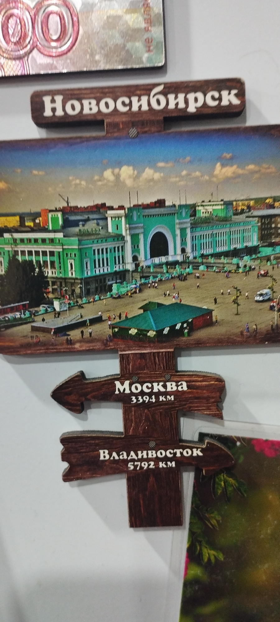 This is a close-up, low-angle, eye-level, indoor photograph of a souvenir refrigerator magnet. The magnet features a depiction of the Novosibirsk train station in Novosibirsk, Russia. Below the train station image are two wooden signposts, each shaped like an arrow. The upper signpost reads "Москва" (Moscow) with "3394 км" (3394 km) underneath. The lower signpost reads "Владивосток" (Vladivostok) with "5792 км" (5792 km) underneath. The overall scene is a still life arrangement, with no people visible. The lighting appears to be artificial, and the background is plain white. The image does not contain any information or visual cues that would place it in the city of Pokosnoye, Russia.