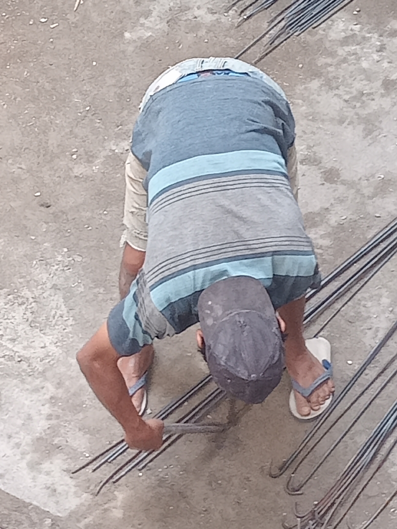 In the city of Simo, Indonesia, a person is seen from a high angle, bent over and working with metal rods on a concrete surface. The individual is wearing a blue and grey striped t-shirt, light-colored shorts, a dark baseball cap, and flip-flops. They are holding a bundle of rebar with their right hand, appearing to be in the process of bending or tying them. The ground is a rough, grey concrete texture, and several long metal rods are laid out around the person, some with hooks at the ends. The scene suggests manual labor in a construction or fabrication setting.