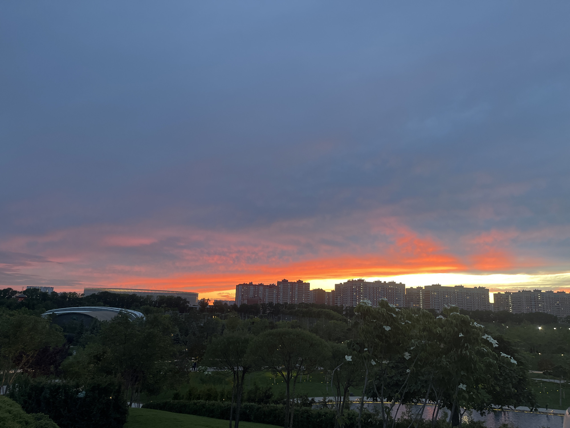 This image captures a vibrant sunset over an urban landscape, likely in Krasnodar, Russia. The sky dominates the upper portion, transitioning from a deep blue-grey at the top to striking bands of orange and red along the horizon, strongly indicating either a sunset or sunrise. Wispy clouds are illuminated by these warm colors, creating a dramatic atmospheric effect.
In the middle ground, a prominent skyline is silhouetted against the bright horizon. This includes several rows of multi-story residential buildings with numerous windows, their dark forms contrasting sharply with the illuminated sky. To the left, a large, modern, circular stadium or arena structure is visible, featuring a light-colored exterior band, possibly the FC Krasnodar Stadium. Closer to the foreground on the left, another modern building with a distinctive curved, sleek roof structure is partially visible amidst the trees.
The foreground is filled with lush greenery, suggesting a well-maintained park or landscaped area. Numerous trees and bushes with varying foliage are visible, some adorned with small white blossoms, adding texture and detail to the scene. A dark, reflective body of water, possibly a pond or canal, can be seen in the lower right, partially obscured by trees. The overall mood is one of serene urban beauty at twilight, with the dramatic sky being the focal point. No discernible text is present in the image.