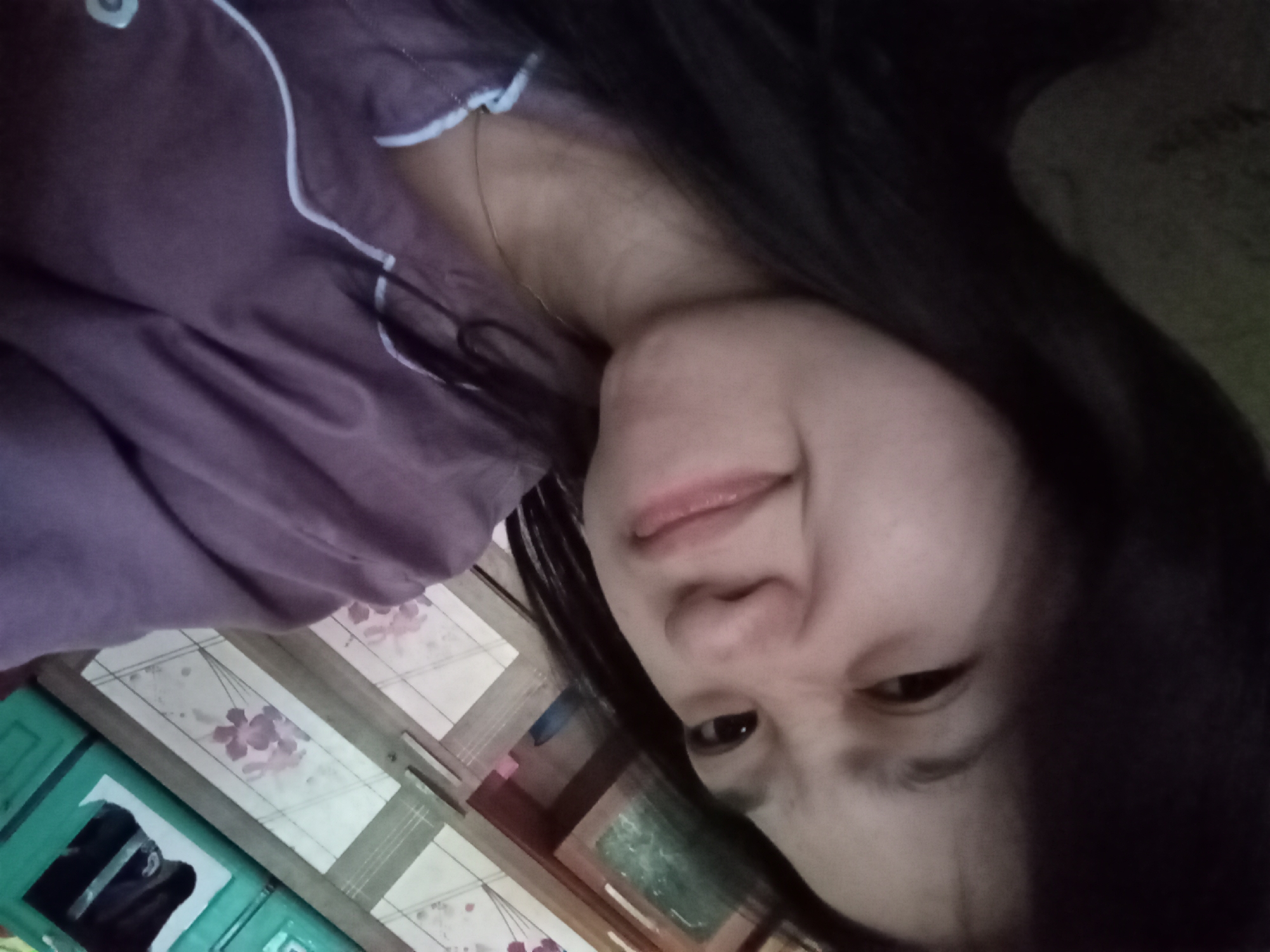 The image is a close-up, selfie-style photograph of a young woman. She has dark hair and is wearing a purple pajama top with white piping. Her facial expression is a playful grimace or smirk, with her eyes squinted and her mouth slightly pursed. The background is out of focus and appears to be an indoor setting, possibly a bedroom, with what looks like furniture or shelves visible. The lighting is dim, contributing to a slightly grainy quality of the image. No specific location in Rejosari, Indonesia, can be identified from the visual information provided.