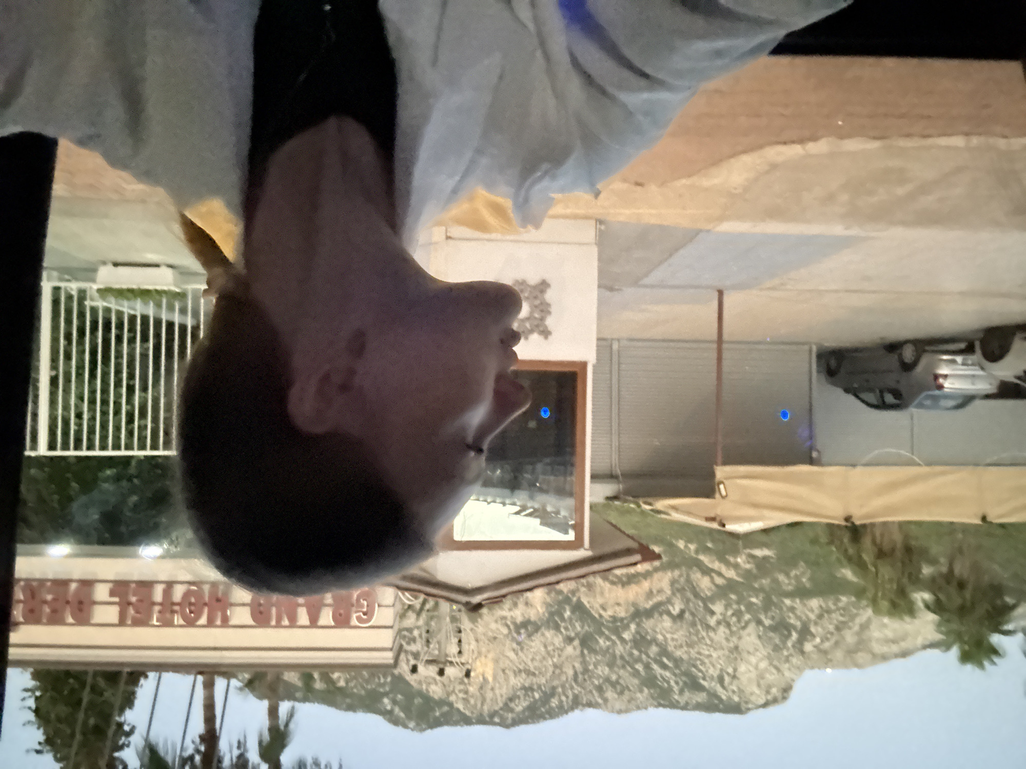 The image shows a young woman from the chest up, her head turned to the right, in profile, looking towards the side. She has light brown hair pulled back into a ponytail. She is wearing a white jacket over a dark shirt. In the background, a sign reads "GRAND HOTEL DEREN", indicating the location in Kemer, Turkey. There are mountains in the distance, palm trees, and some buildings with awnings. A silver car is parked to the left. The overall scene appears to be outdoors, possibly near a hotel or resort.