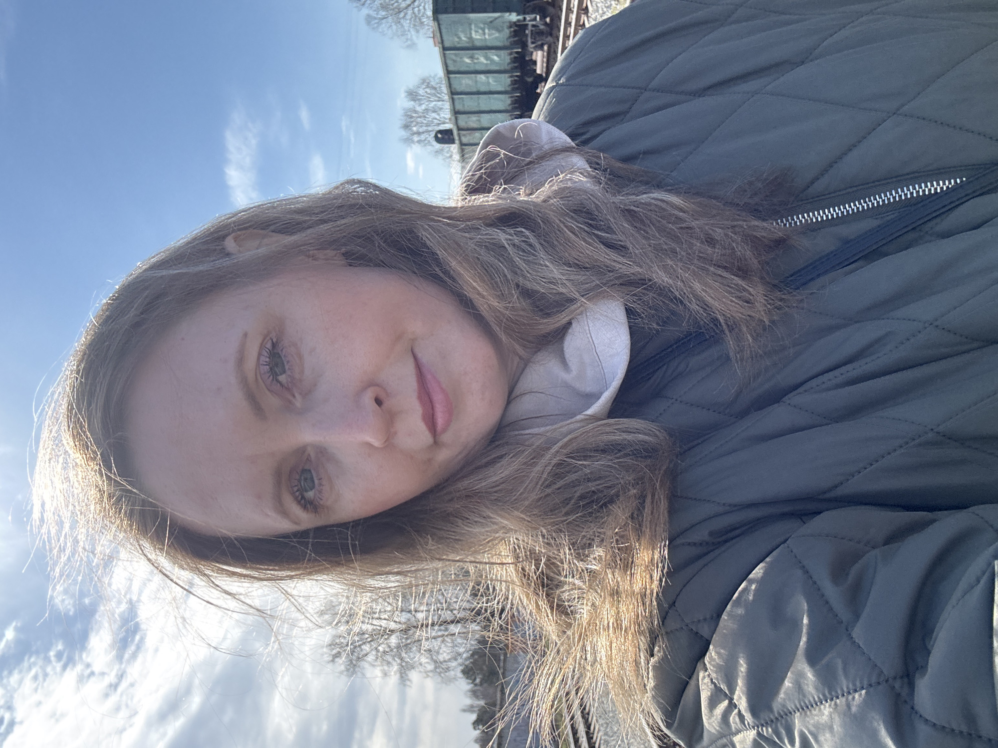 A woman with long blonde/brown hair is taking a selfie outdoors on a sunny day. She is wearing a quilted green jacket and a light-colored hoodie. Behind her, a train and train tracks are visible, along with bare trees and a blue sky with wispy clouds. The location may be Chaikovsky, Russia, given the instructions.