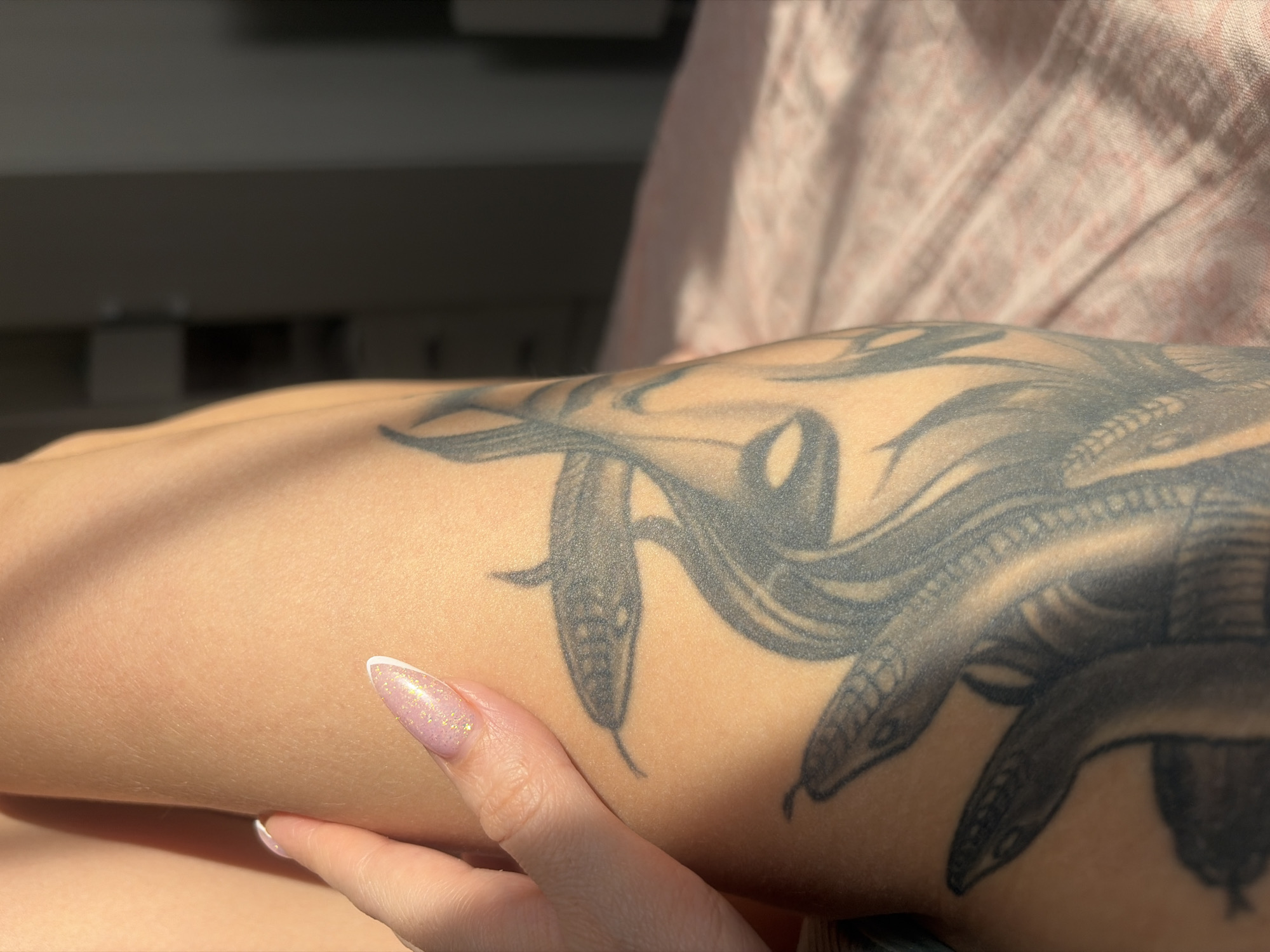 The image depicts a close-up of a person's arm with a detailed black and grey tattoo of snakes, possibly Medusa. The arm is fair-skinned and a hand with decorated fingernails is also visible. The background is blurred, showing light colors suggesting a room interior. The location in the city of Saint Petersburg, Russia is not discernible from the image.