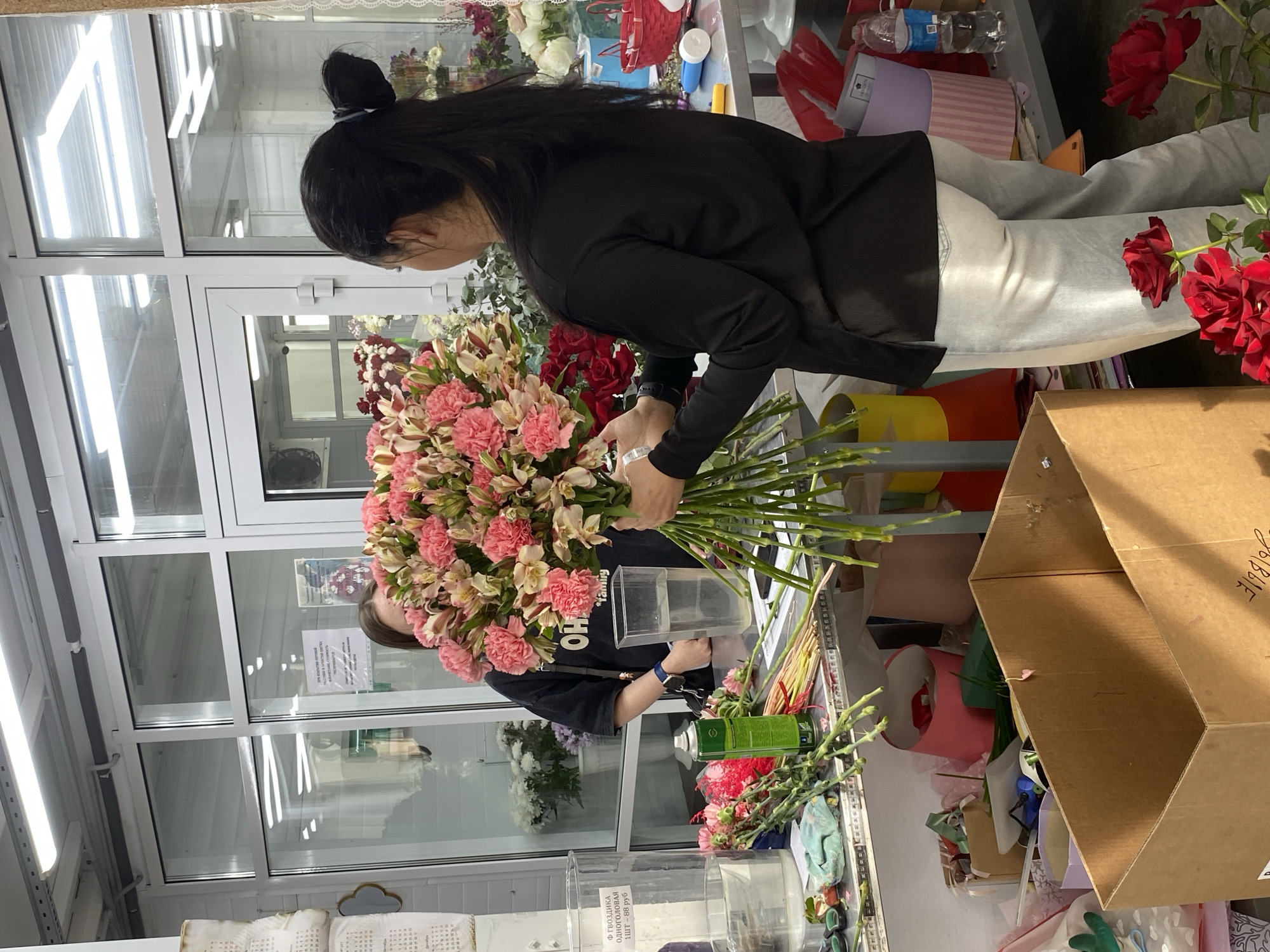 A florist is arranging flowers in a flower shop. The florist is a woman with long black hair pulled back in a bun. She is wearing a black long-sleeved shirt and light-colored pants. She is holding a large bouquet of pink and white alstroemeria flowers. The flowers are arranged on a table with other flowers and floral supplies. In the background, there is another person in a black t-shirt. The shop has a large window with a view of the city of Yekaterinburg, Russia. A paper with Russian writing is visible. There are numerous flower arrangements and floral supplies scattered on the table and shelves. A cardboard box is visible on the right side of the frame.