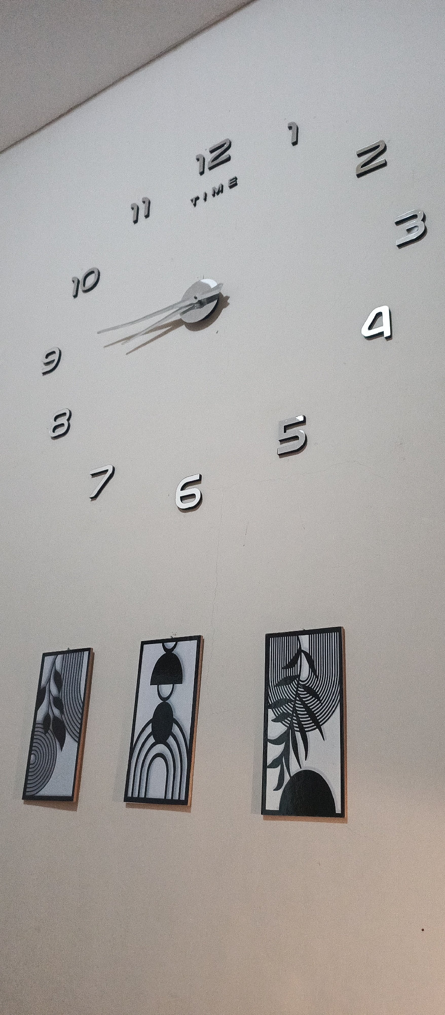 The image shows a modern wall clock with silver numbers mounted on a cream-colored wall. The clock has a central mechanism with two silver hands, the hour hand pointing between the 10 and 11, and the minute hand pointing towards the 2. The numbers 1 through 12 are visible around the clock face, though some are obscured by the angle of the shot. Below the clock, three framed abstract art pieces are hung in a horizontal line. Each frame contains a black and white graphic design. The leftmost frame features a swirling pattern with leaf-like shapes. The middle frame has a series of concentric arcs and a rounded black shape at the top. The rightmost frame displays a stylized leaf design against a background of curved lines. The lighting suggests an indoor setting, with a soft light source illuminating the wall and objects. The image is taken from a slightly elevated angle looking down towards the art pieces.