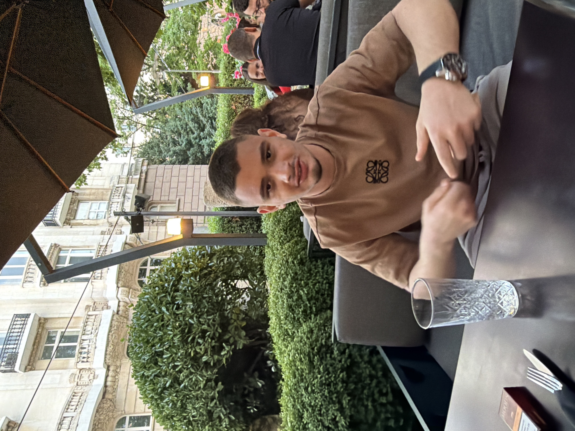 This image captures a man seated at an outdoor cafe or restaurant in Sofia, Bulgaria, looking directly at the camera. The photo is rotated 90 degrees counter-clockwise from its natural orientation.
The primary subject is a young man with short dark hair and light stubble, wearing a plain brown long-sleeved shirt. A distinct black Loewe Anagram logo is visible on the left chest of his shirt. He is wearing a dark-strapped watch on his left wrist and has his hands resting on the dark table in front of him. He appears to be smiling slightly, suggesting a relaxed and friendly atmosphere.
On the table in front of him, there is a clear glass with an ornate, diamond-patterned design, containing liquid. Next to it, a fork and knife are neatly placed, along with a red and white cigarette pack that clearly shows the brand name "MOODS."
In the background, other patrons are visible, seated at tables, suggesting a busy dining environment. Lush green foliage, including bushes and trees, creates a natural barrier or garden-like ambiance around the seating area. Above, large dark umbrellas provide shade. Beyond the greenery, ornate, light-colored buildings with intricate architectural details and balconies are visible, typical of older European city architecture. Street lights are also visible, one of which is illuminated, hinting at either a late afternoon/early evening time or an overcast day. The overall impression is a pleasant, social outdoor setting.