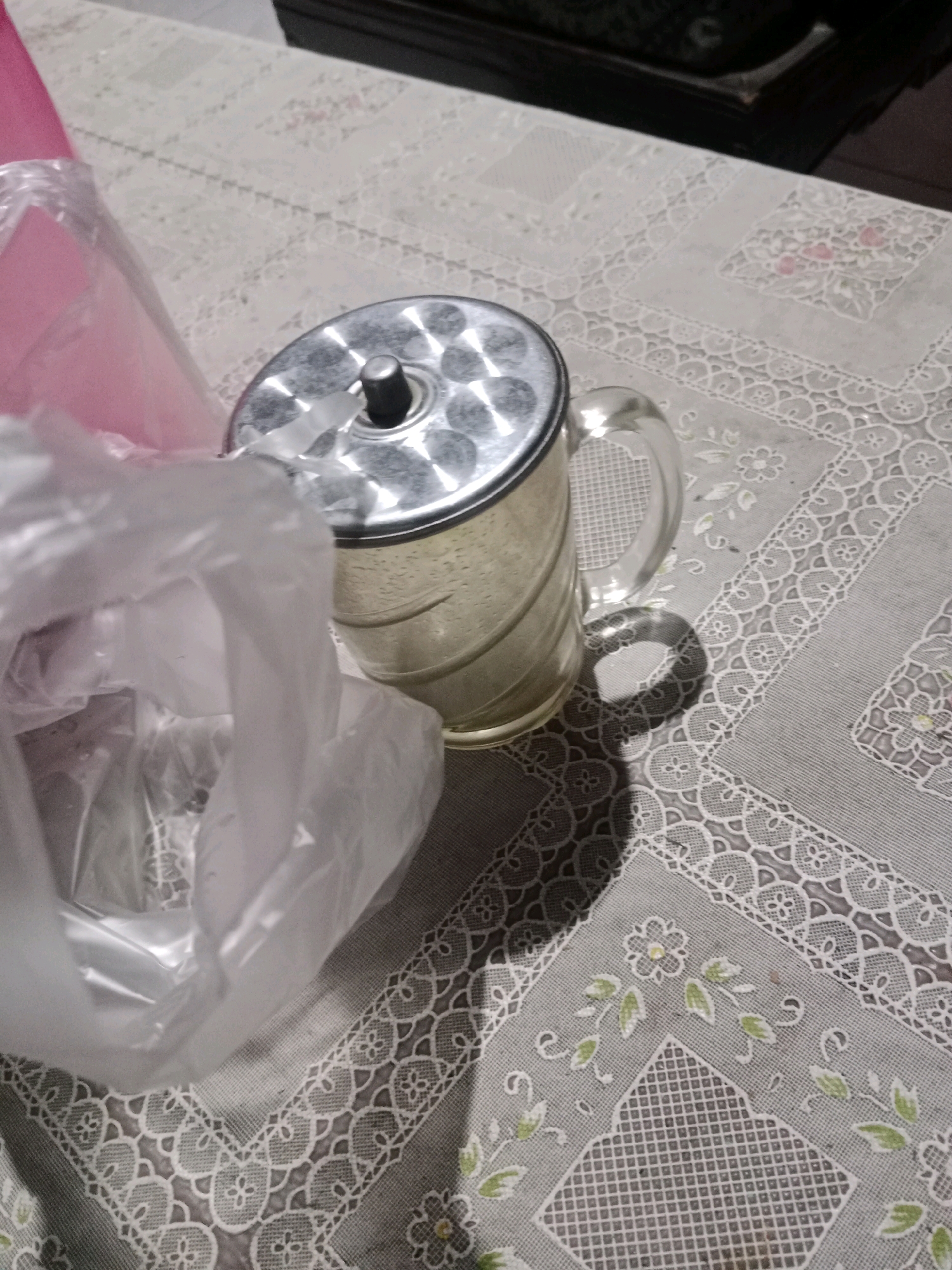 A stainless steel mug with a glass handle sits on a tablecloth with floral and geometric patterns. A white plastic bag is partially obscuring the mug. The setting appears to be indoors, on a table. There is no indication of Ringinrejo, Indonesia.