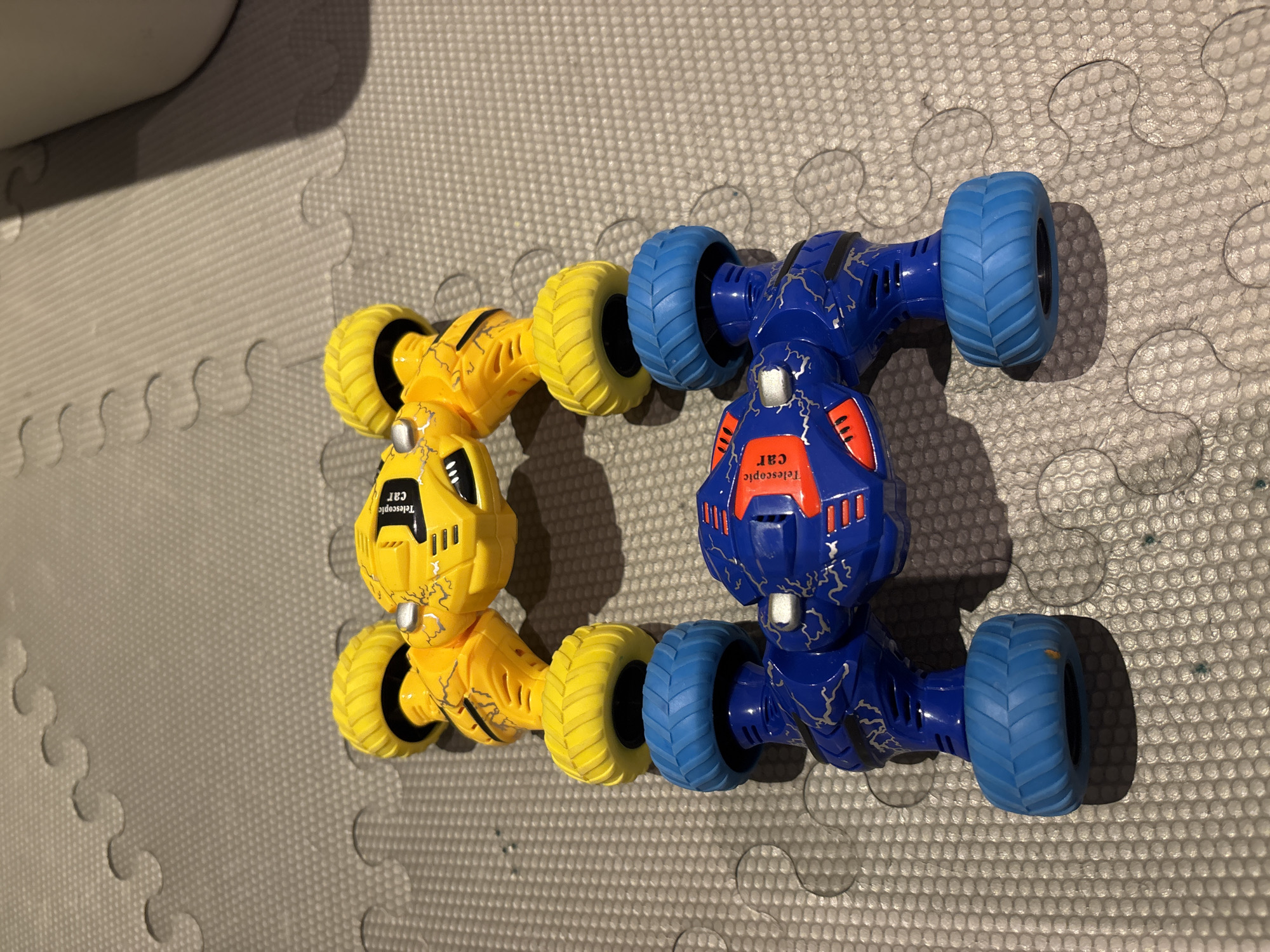 A yellow and blue toy car, designed to resemble a robot or monster, is positioned vertically on a gray, textured floor mat. The car has four wheels, two yellow and two blue, and is split into two main sections. The top section is yellow with black markings resembling lightning and has two yellow wheels. The bottom section is blue with red and black accents and also has two blue wheels. The car is not in motion and there are no people or other objects in the immediate vicinity. The lighting suggests an indoor environment. There is no information in the image that would indicate the location within Dubai, United Arab Emirates.
