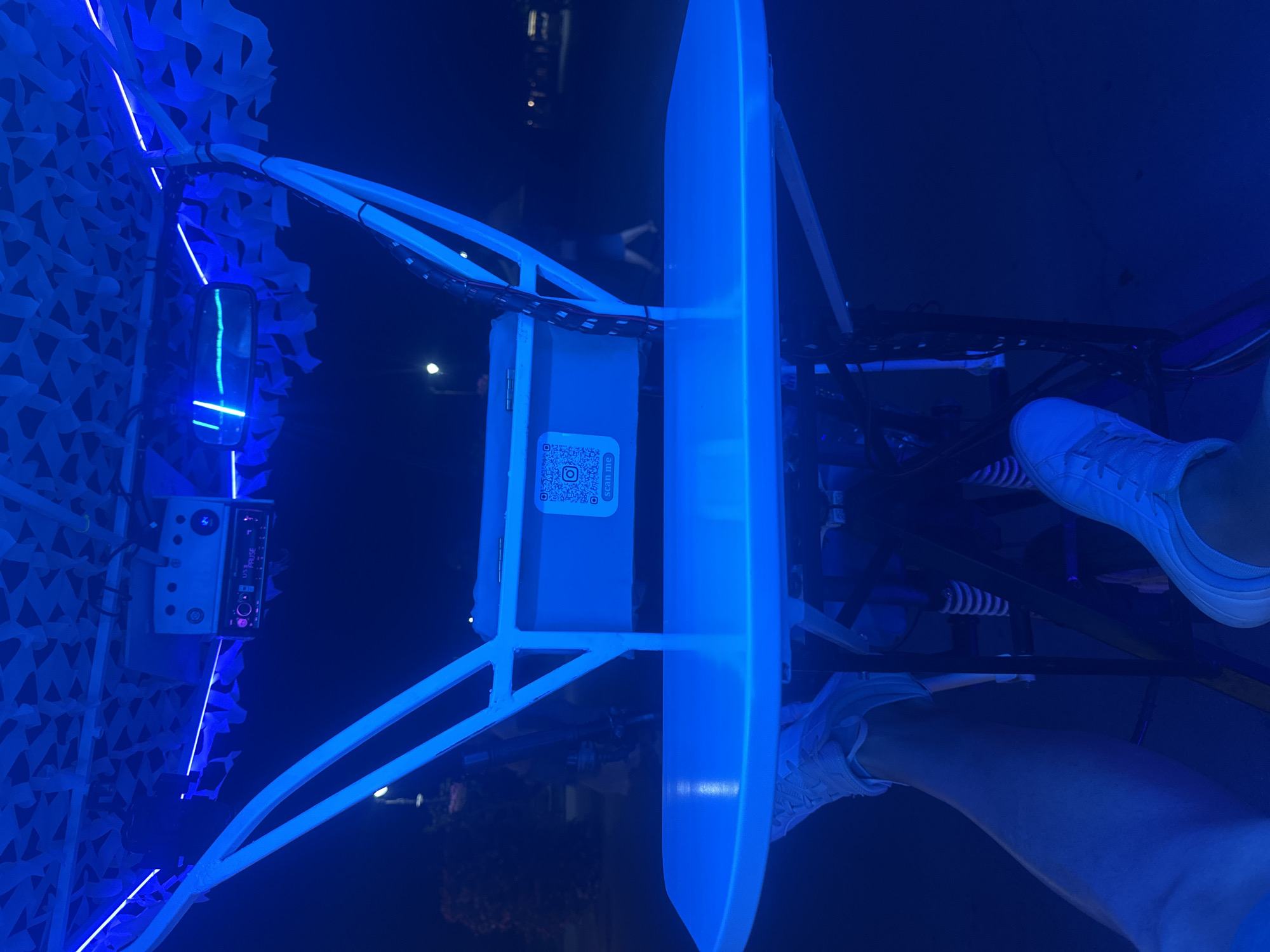 The image is an indoor, low-angle, first-person perspective shot of a person's legs and feet, wearing white sneakers, on what appears to be a modified vehicle at night. The scene is illuminated by blue LED lights. The vehicle has a white frame and a white tabletop in the foreground. Mounted above the tabletop is a car stereo and what looks like a rear-view mirror. A camouflage-patterned fabric hangs from the ceiling structure. A QR code is visible on a surface behind the tabletop. The background is dark, suggesting an outdoor setting at night, possibly a parking lot or street, with blurred lights and figures of people in the distance. There are no discernible landmarks or clues to specify the location within Varna, Bulgaria.