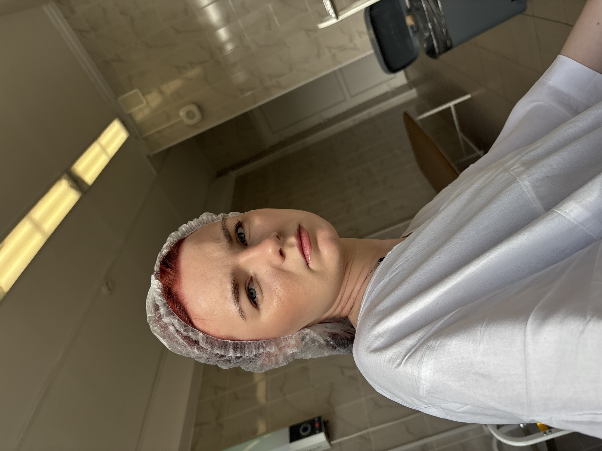 A young woman with red hair, wearing a disposable shower cap and a white medical gown, looks at the camera with a serious expression. She appears to be in a brightly lit room with tiled walls and fluorescent lighting, possibly a medical or salon setting. In the background, a trash can and a table with metal legs are visible. The overall impression is one of a clinical or preparatory environment. The scene does not provide enough information to determine a specific location within Saint Petersburg, Russia.