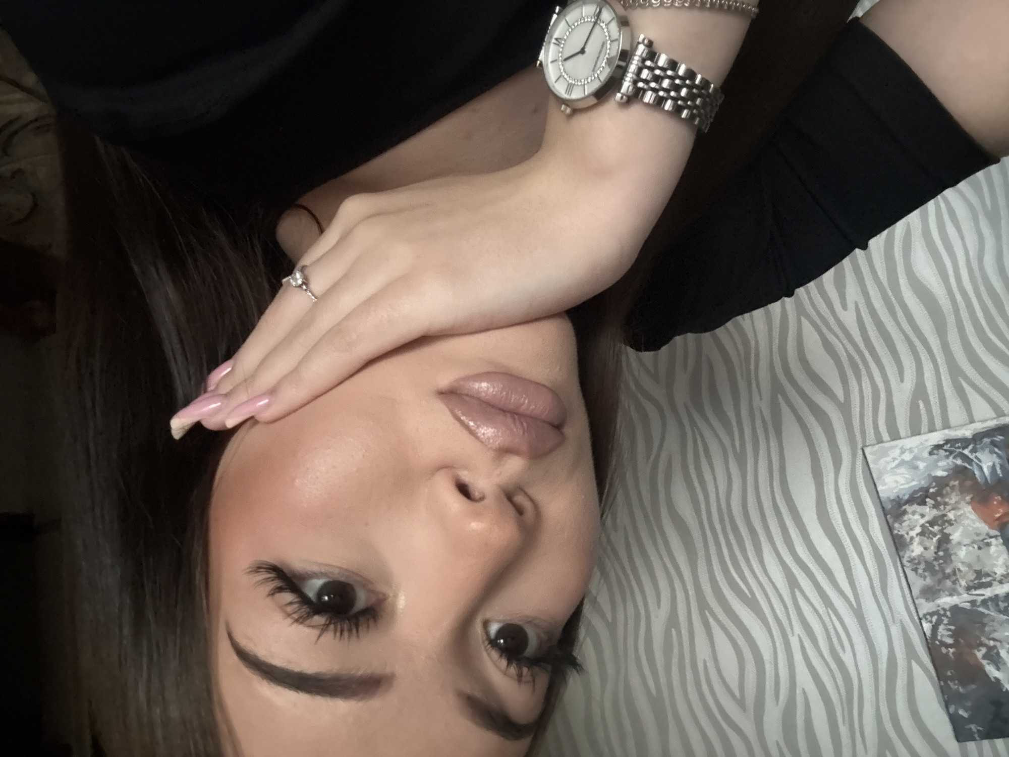 A young woman is lying on a bed or other flat surface covered in zebra-striped bedding. She is wearing a black top, a silver watch, a bracelet and a ring. She has brown hair and full makeup. The photo appears to be taken in a room with a painting on the wall. There is no indication of Naberezhnye Chelny, Russia.
