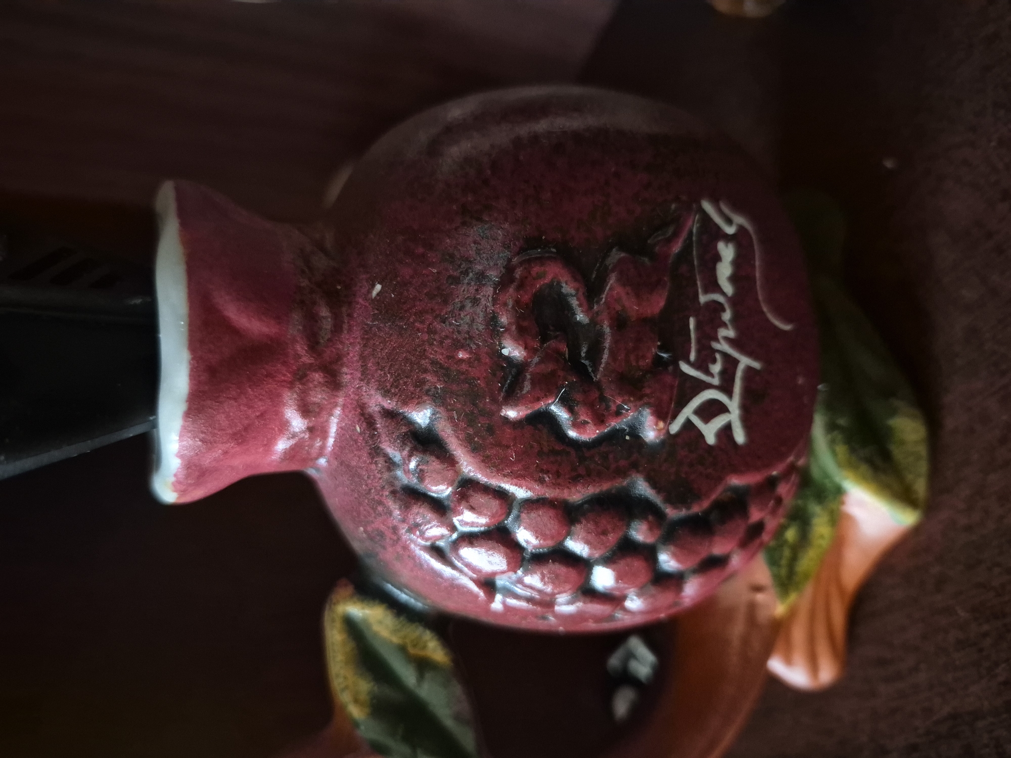 The image presents a close-up view of a small, decorative ceramic or clay vessel. The vessel is primarily a mottled dark red or burgundy color with darker accents, displaying a textured, somewhat rough surface, possibly glazed. It has a rounded, bulbous body that tapers upwards to a distinct white rim at its opening.
On one side, the vessel features raised relief ornamentation resembling clusters of grapes or berries. On an adjacent side, there is a more abstract, swirling raised pattern. Near the base, a white inscription or signature in a stylized script, possibly Armenian, is visible, appearing to spell characters such as 'Դapwag' or similar. A dark, thin, and somewhat rectangular object is inserted vertically into the white-rimmed opening at the top.
The vessel rests on a dark, indistinct surface, possibly fabric. Partial views of sculpted, leaf-like elements are visible at the base: one in an orange hue with prominent ridges, and another in dark green with yellow highlights. The background is dark and heavily out of focus, indicating an indoor setting with subdued lighting. The visual content of the image does not provide any information regarding a specific geographical location, such as Vanadzor, Armenia.