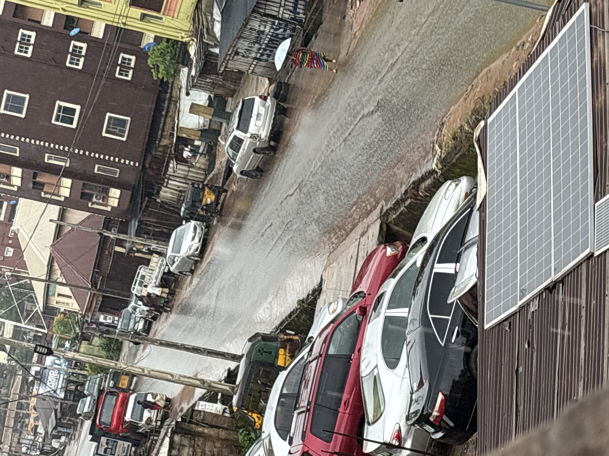 A street scene in Nkpor, Nigeria, during a heavy rainfall shows a variety of parked vehicles, including cars, trucks, and a tricycle, lining a wet, muddy road. Buildings of varying heights and architectural styles are visible on either side of the street, some appearing to be commercial establishments with signs. People are present, with one individual holding an umbrella and walking on the road, while others stand near parked vehicles. The overall atmosphere suggests an active urban environment experiencing inclement weather.
