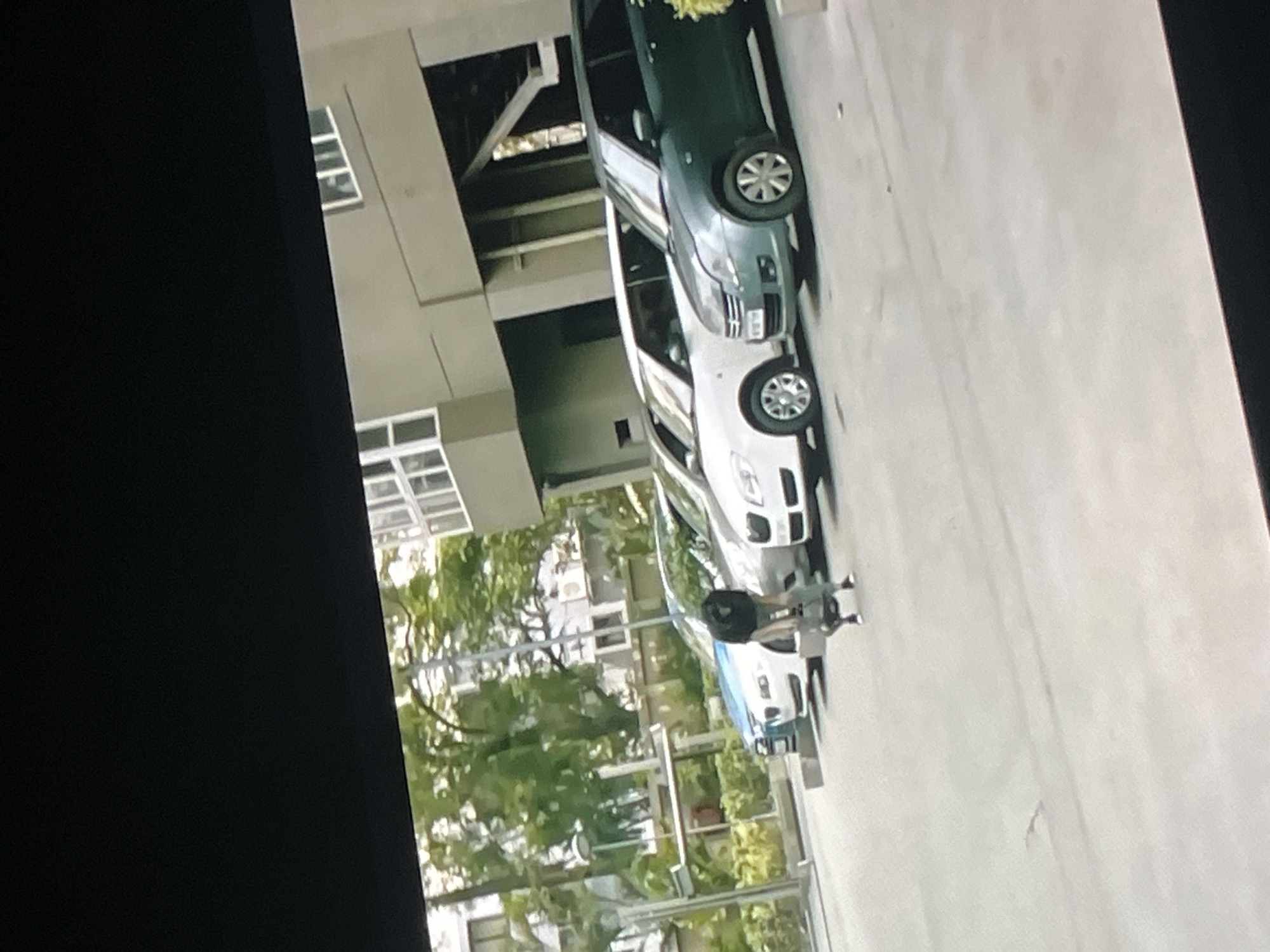 This is a vertical, slightly blurry, eye-level shot taken outdoors in what appears to be an urban or suburban setting. The scene is dominated by a light-colored, possibly concrete, surface, likely a road or pathway, with a darker area on the left side that could be a shadow or a different surface.
There are two cars visible. The car in the foreground, on the right, is white. Behind it is a dark green or black car. Both cars appear to be parked. A person is visible between the two cars, appearing to be a woman wearing dark clothing and possibly pushing or interacting with something. The person is in motion, with their legs slightly blurred.
The background shows architectural elements, suggesting buildings, with windows visible. There are also trees with green foliage, indicating a potentially leafy environment. The lighting suggests it is daytime, possibly late afternoon given the angle of any potential shadows, though this is difficult to determine with certainty due to the image quality. The weather appears to be clear and dry. No text is discernible in the image. The overall impression is a casual, everyday scene, possibly in a residential or commercial area.