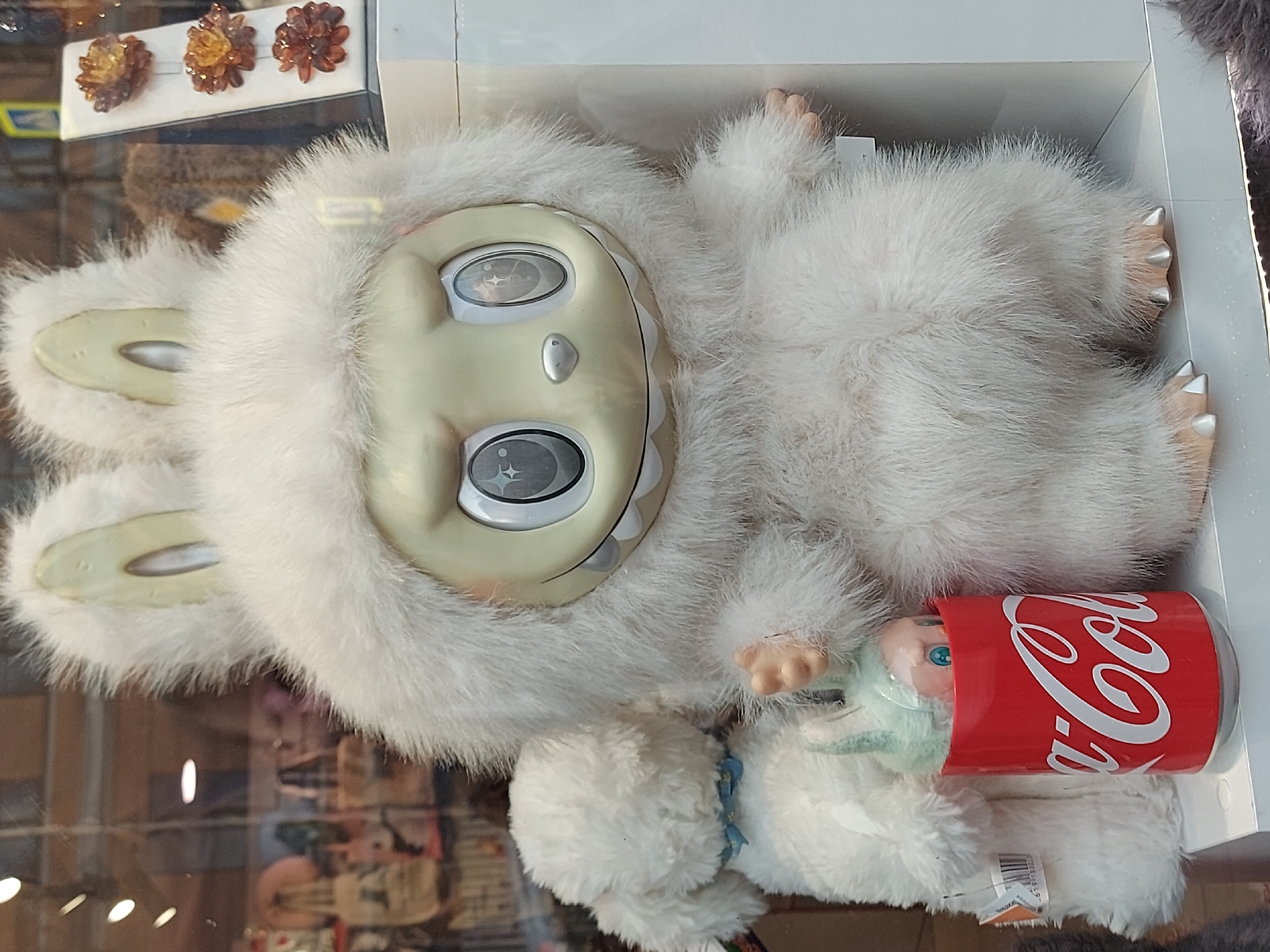 The image shows a furry, white, rabbit-like doll with a pale face, large eyes, and a toothy grin. The doll has two long ears and is holding a small doll wrapped in a blue cloth, with only its face visible, next to a red Coca-Cola can. The doll is situated inside a glass display case, with other items and reflections visible in the glass. In the upper left corner of the frame, three decorative, amber-colored flower-like ornaments are affixed to the back of the case. The location is Saint Petersburg, Russia.