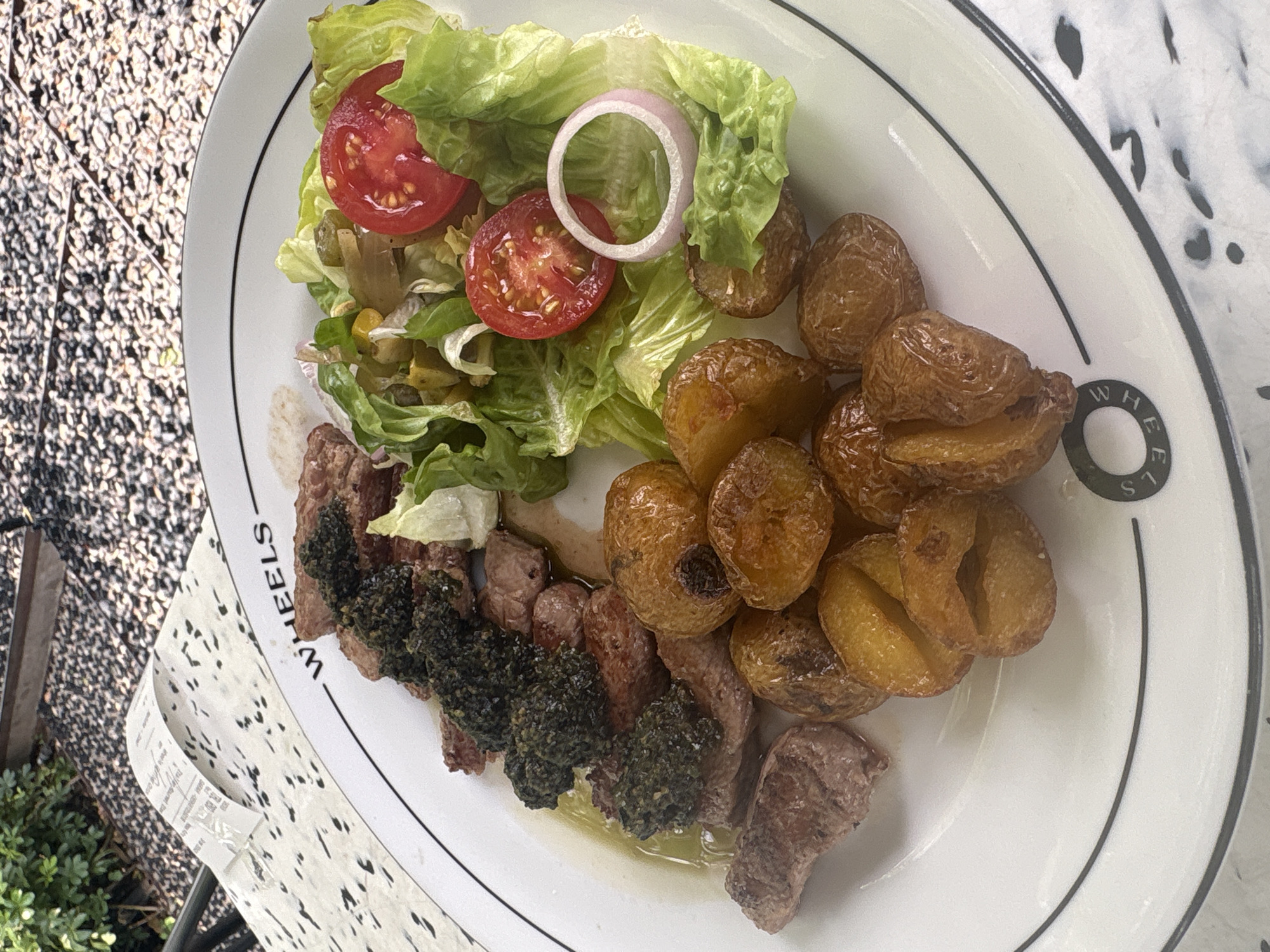 A close-up, high-angle shot features a plate of food on a table, suggesting an outdoor dining setting. The plate, which has "WHEELS" printed on its rim, contains a meal consisting of sliced steak topped with a dark green sauce, small roasted potatoes, and a side salad with lettuce, cherry tomatoes, and a slice of red onion. A receipt is visible on the patterned tablecloth in the background, which appears to be outdoors. The lighting suggests it is daytime. The scene does not provide any specific visual cues to determine its location within Bandung City, Indonesia.
