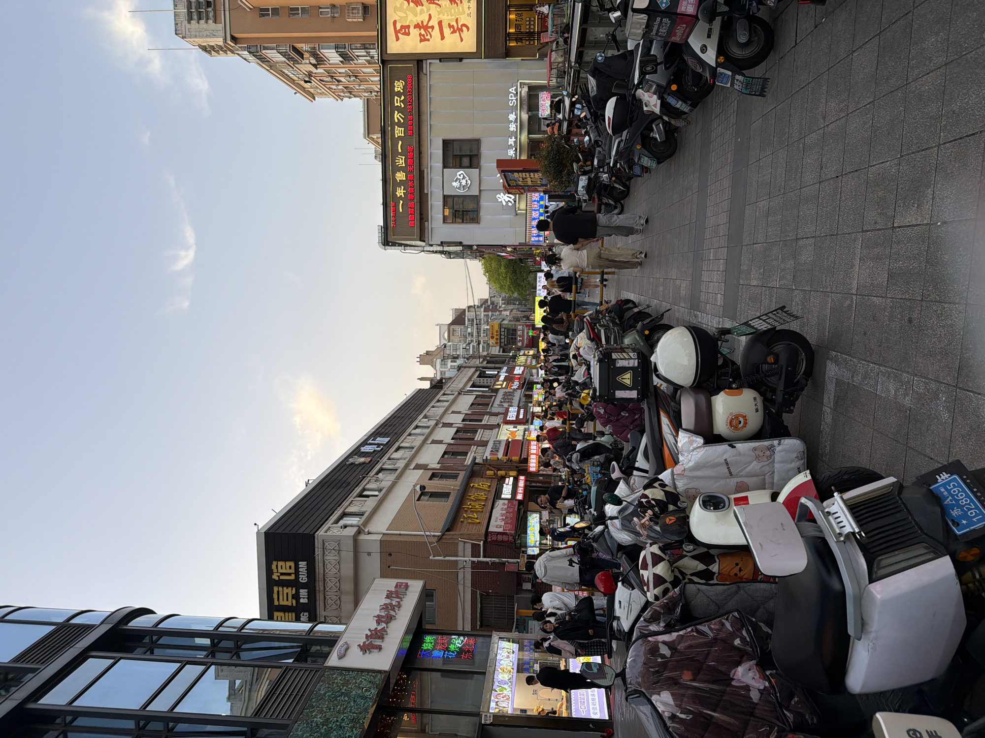 The image captures a bustling street scene in Jiangning District, China, filled with parked electric scooters and pedestrians. Buildings with illuminated signs in Chinese and English are lined along the street. One sign reads "BIG BIN GUAN" in gold letters, another advertises "Domino's Pizza". A crowded area of people is visible in the middle distance, suggesting a busy thoroughfare. The street pavement consists of grey square stones. The sky above is visible and light blue.