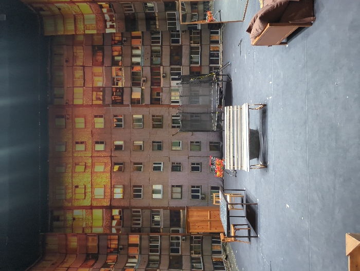 The image displays a theatrical stage set designed to represent a residential courtyard in Moscow, Russia. The backdrop is a large, curved, painted representation of a multi-story, grey, weathered apartment building, characteristic of Soviet-era architecture, with numerous windows. Many of these windows, particularly on the upper floors and right side, emit a warm, orange-yellow light, simulating interior illumination at night or dusk. The stage floor is a flat, dark grey surface with some minor white markings.
Arranged on the stage are several objects: On the left, a dark rectangular table with a light, speckled surface is flanked by two simple wooden chairs. Behind the table stands a tall, light brown wooden structure, possibly a door or shed facade. In the center, a worn, light-colored wooden park bench with a rusted frame is positioned in front of a dark, low, circular object, possibly a planter. Further back on the left, a small cart or stand holds a cluster of artificial red and orange flowers. To the right of the bench, a large, circular trampoline with a black mat and surrounding netting stands on a metal frame. In the far right foreground, a portion of a brown sofa with dark cushions is visible. On the far right, a large, rectangular mirror with a dark frame stands upright, angled slightly into the scene. No individuals are present.