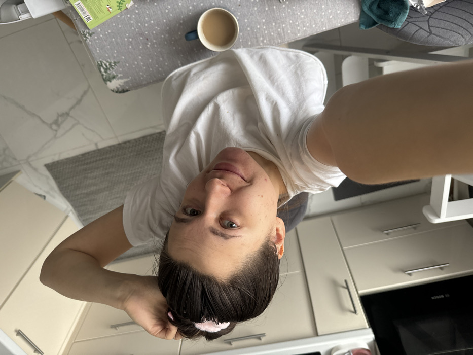 An overhead-angle selfie captures an adult female within what appears to be a residential kitchen or dining area. The exact location in Odintsovo, Russia, is not discernible from the visual evidence. The female, positioned centrally, has dark hair pulled back with a light pink scrunchie. She wears a plain white short-sleeved t-shirt. Her right arm extends towards the camera, while her left hand is near the back of her head. Her face is visible, with eyes looking towards the camera and a slight smile. The background on the left reveals light-colored kitchen cabinets with metal handles and a dark, built-in appliance displaying a faint "BOSCH" logo. The floor consists of large white tiles with grey veining, partially covered by a grey textured rug. To the right, a table is visible, draped with a light grey tablecloth featuring a white evergreen tree pattern. A blue mug containing a light brown liquid is on the table, alongside a green object, possibly packaging or a book. A white chair and part of another chair or stool with a grey cushion are also visible. A dark teal cloth rests on an unseen object in the lower right corner.