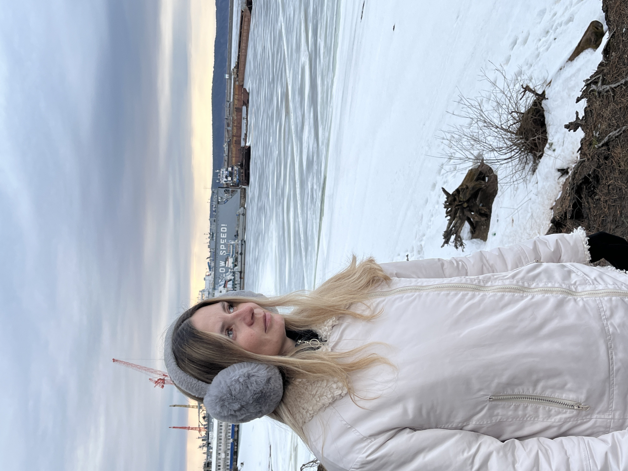 The photograph depicts a woman outdoors in winter. She is wearing a light-colored jacket and gray earmuffs. Behind her is a landscape covered in snow, a body of water, and industrial buildings, including cranes. A sign with the words "LOW SPEED!" is visible on one of the buildings.
Based on the provided location information, the scene is likely in or around the city of Chaikovsky, Russia. The industrial buildings and body of water suggest a riverside location or a port area within the city.