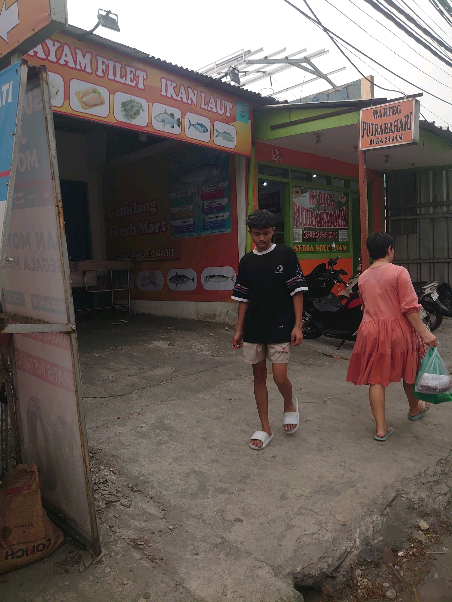 In Depok, Indonesia, two individuals walk on a paved outdoor area. In the foreground, a young person wearing a black t-shirt with white and red accents on the sleeves and tan shorts walks from left to right, holding a cigarette in their right hand. They are wearing white slides. To their right, a person in a coral-colored tiered dress walks in the same direction, carrying a green plastic bag filled with items in their right hand. They are wearing blue slides.
Behind the individuals, there are two businesses. The one on the left has a large orange and yellow banner that reads "AYAM FILET IKAN LAUT" (Chicken Fillet Fish Sea) with illustrations of chicken and various fish. Below this banner, a sign reads "Gemilang Fresh Mart Grosir & Eceran" (Gemilang Fresh Mart Wholesale & Retail). The business on the right has a prominent orange sign with black text stating "WARTEG PUTRABAHARI BUKA 24 JAM" (Warteg Putrabahari Open 24 Hours) and "SEDIA SOTO AYAM" (Chicken Soto Available). Both establishments appear to be food vendors.
The environment is somewhat disheveled, with some debris and cracks visible on the concrete ground. Electrical wires are visible overhead. Motorcycles are parked near the establishments. The overall impression is of a commercial street scene in a moderately urban Indonesian setting.