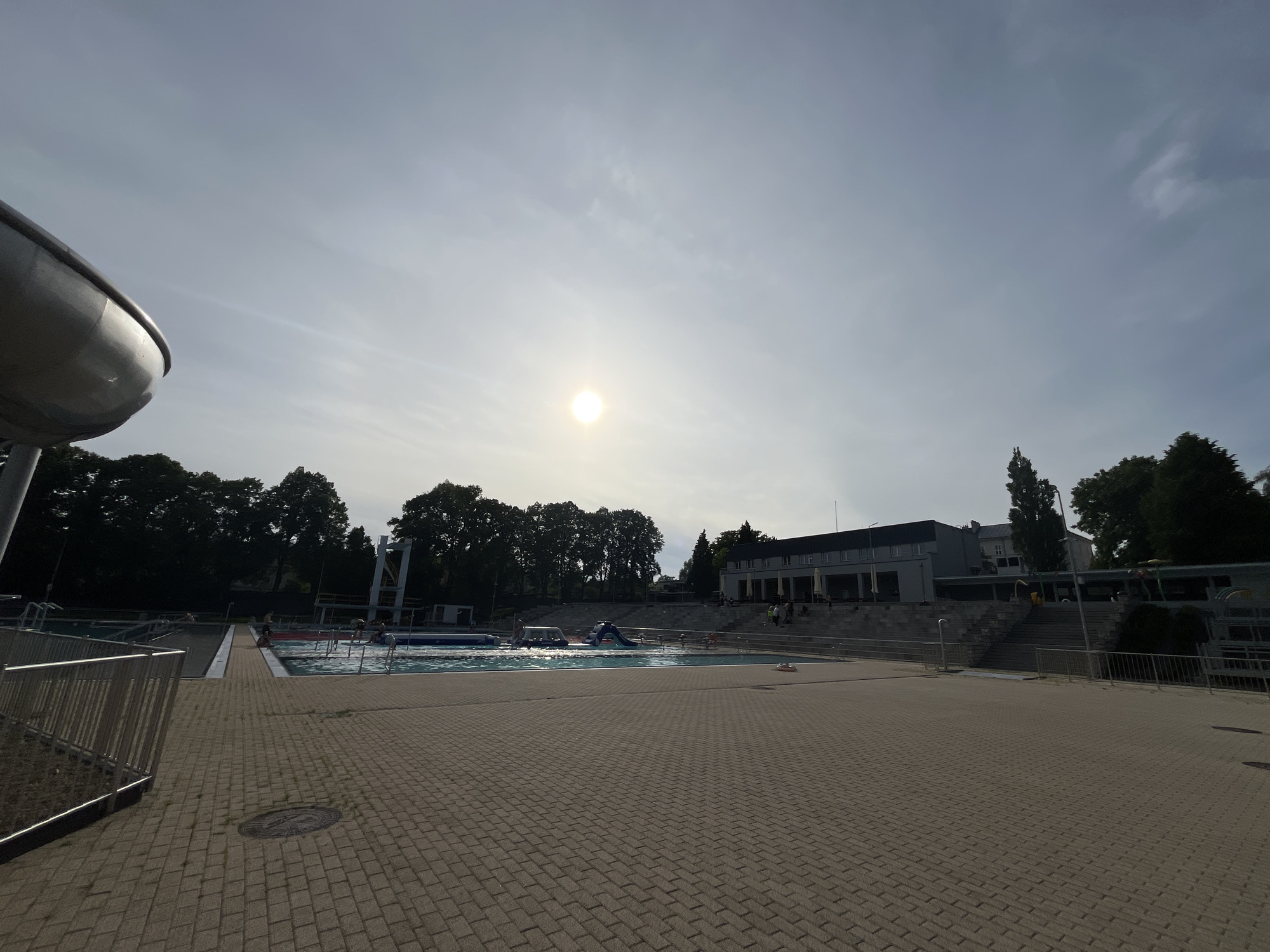 This wide-angle shot captures an outdoor swimming pool complex under a bright, hazy sky. The sun is prominent and centrally located, causing a significant lens flare and backlighting much of the scene.
In the foreground, a large expanse of light-colored, interlocking pavers dominates the lower half of the frame. To the left, a curved, silver-toned water slide structure extends into the frame from the top left corner. Metal railings border parts of the paved area. A circular manhole cover is visible on the ground.
Beyond the paved area, at least two swimming pools are visible. The main pool in the midground contains several blue and white inflatable play structures, and a few people can be seen in the water and on the pool deck, appearing small and indistinct due to the distance and lighting. To the far left, another section of a pool or water area is partially visible, with a tall white diving platform in the background.
The background features a long, two-story building with a grey roof and light-colored walls, possibly housing changing rooms or a restaurant, with outdoor seating visible on its elevated terrace. Concrete stairs lead up to this building. A dense line of dark-green trees runs along the entire background behind the pools and building, forming a natural barrier.
The scene depicts a leisurely summer day at a public swimming facility, with people enjoying the water and sun. The strong backlighting makes the foreground elements appear somewhat shadowed, contrasting with the bright sky. No visible text can be clearly identified.