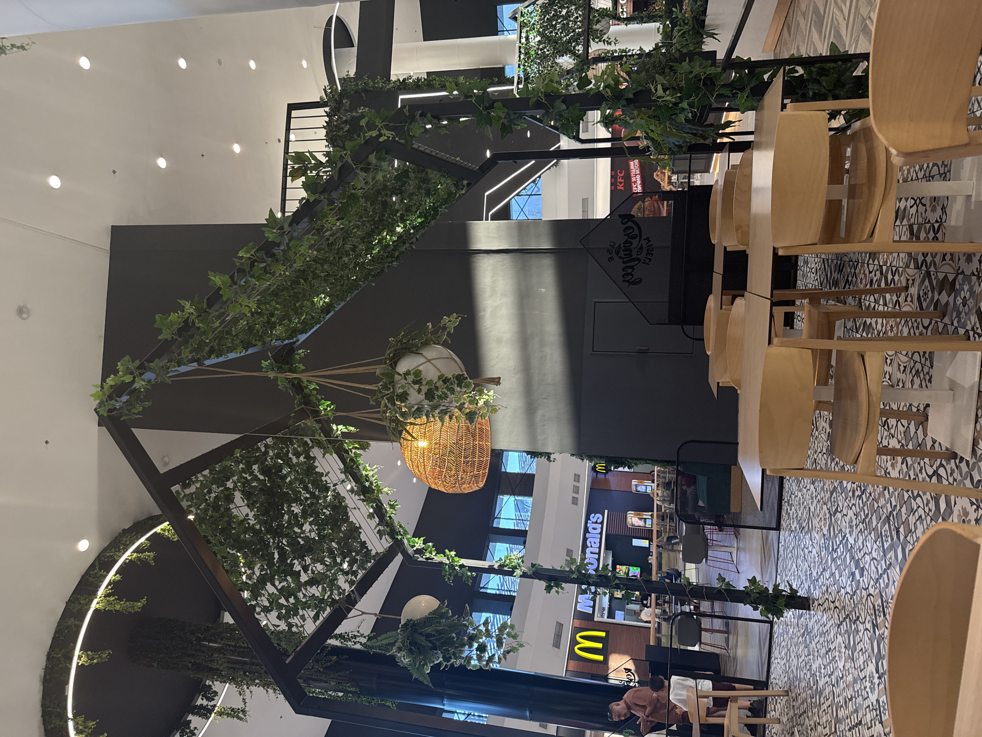 The image depicts an indoor seating area in a modern mall, specifically at the "Mall Galleria" in Stara Zagora, Bulgaria. In the foreground, several light-colored wooden tables and chairs are arranged on a patterned tiled floor. A black metal structure, adorned with artificial green ivy, frames the dining area. Suspended from this structure are two hanging planters, one containing a woven basket with a light bulb, and another white planter also with greenery. In the background, a McDonald's restaurant is visible with its distinctive yellow "M" logo and signage. Further to the right, a KFC restaurant with its logo is also present. People are sparsely scattered throughout the background, some appearing to be waiting or dining. The overall ambiance suggests a contemporary and somewhat nature-inspired food court or dining space within a shopping complex.