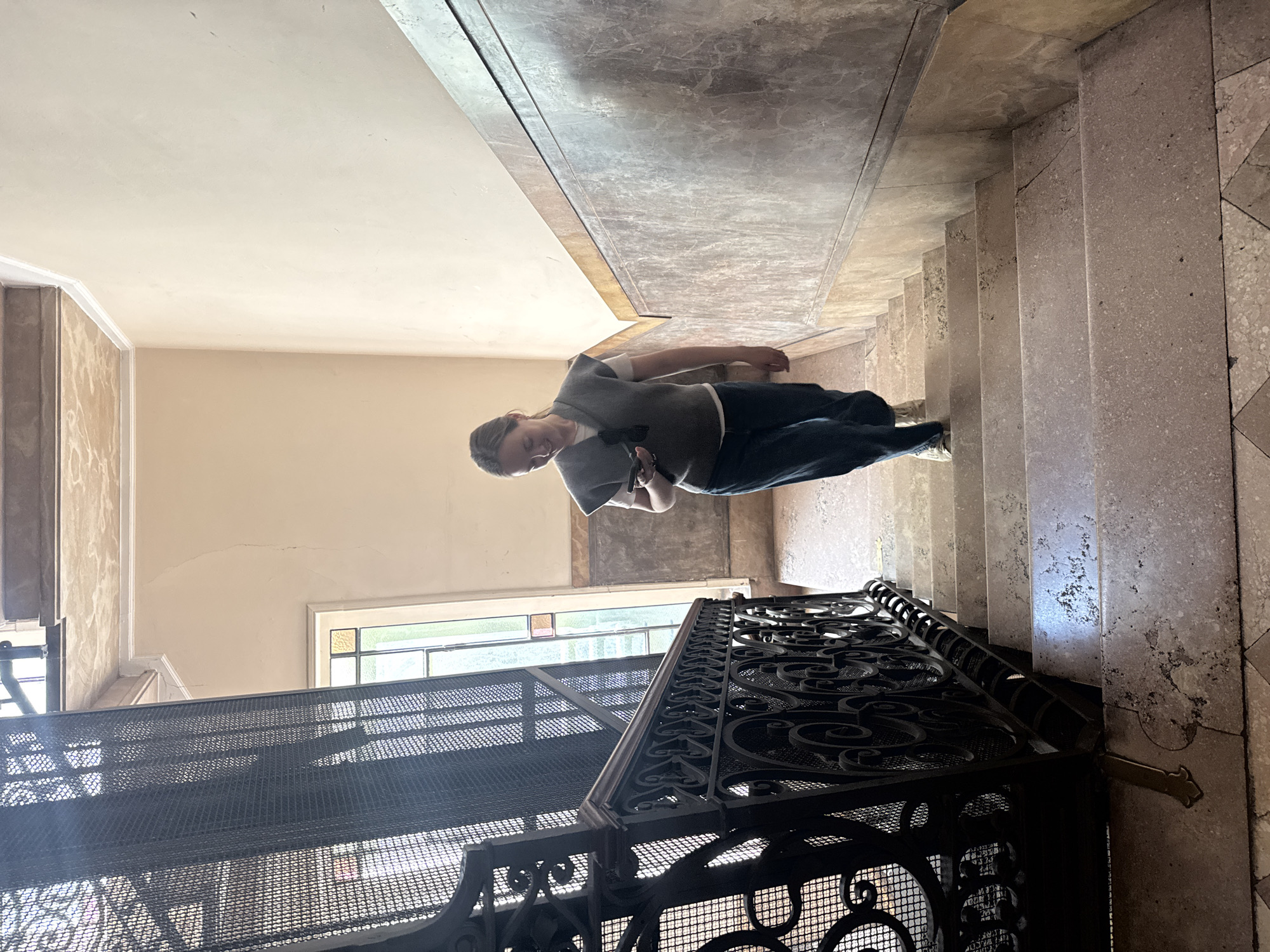 A person is standing on a landing in a staircase in Milan, Italy. The individual, wearing a gray top and dark pants, is holding a small, dark object in their hands. The staircase features marble steps and walls, and is adjacent to a black wrought iron lift cage with ornate decorative designs. A window is visible in the background, providing light to the interior. The lift has a mesh enclosure. The walls are painted in light shades.