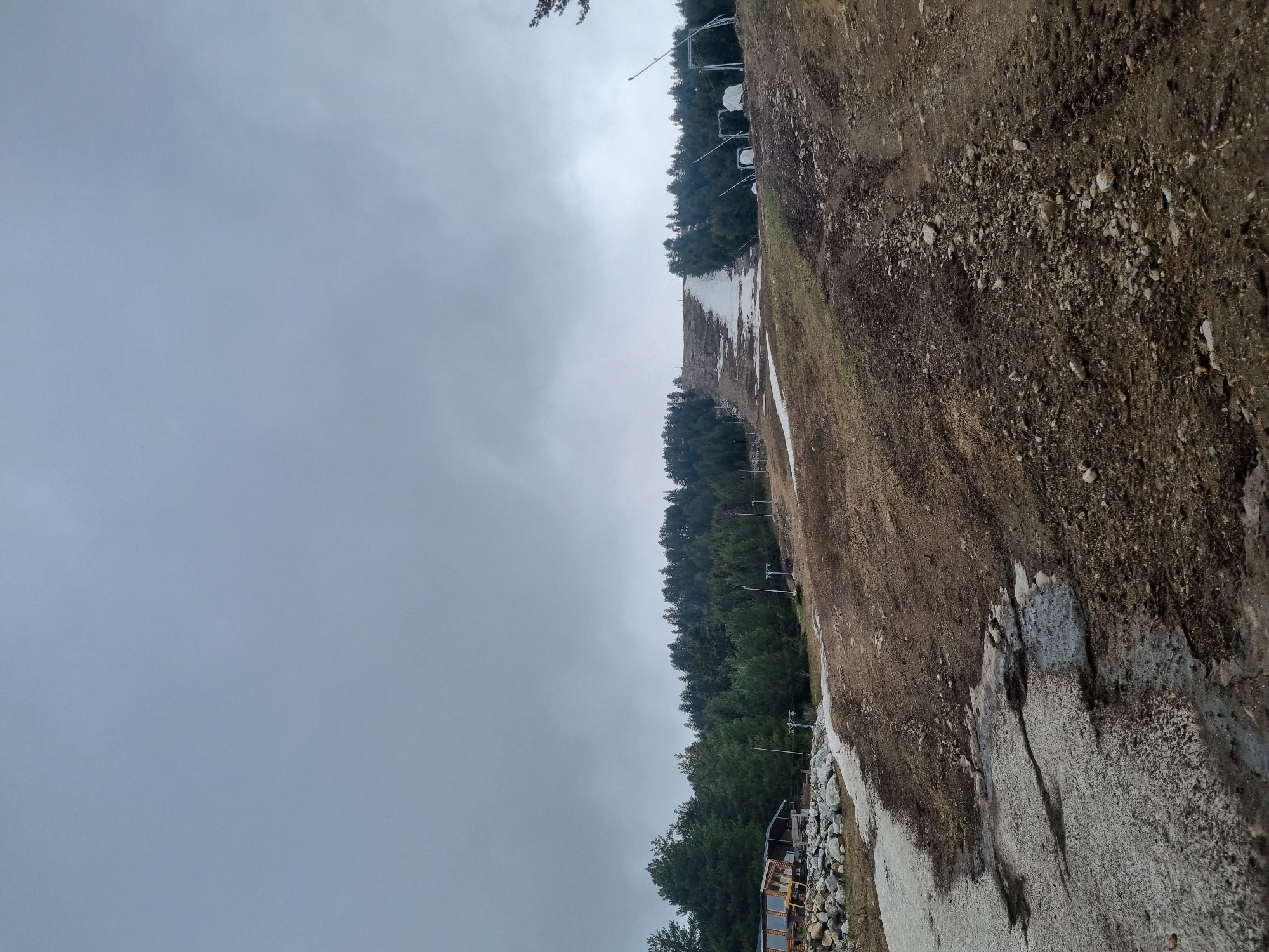 The image depicts a ski slope in Govedartsi, Bulgaria, characterized by sparse snow cover. The slope is bordered by dense coniferous trees on one side and exposed soil/gravel on the other. Ski lift infrastructure is visible along the edge of the tree line. The sky is overcast, suggesting poor weather conditions. The lower portion of the image includes an accumulation of rocks and what appears to be a structure. No people are visible.