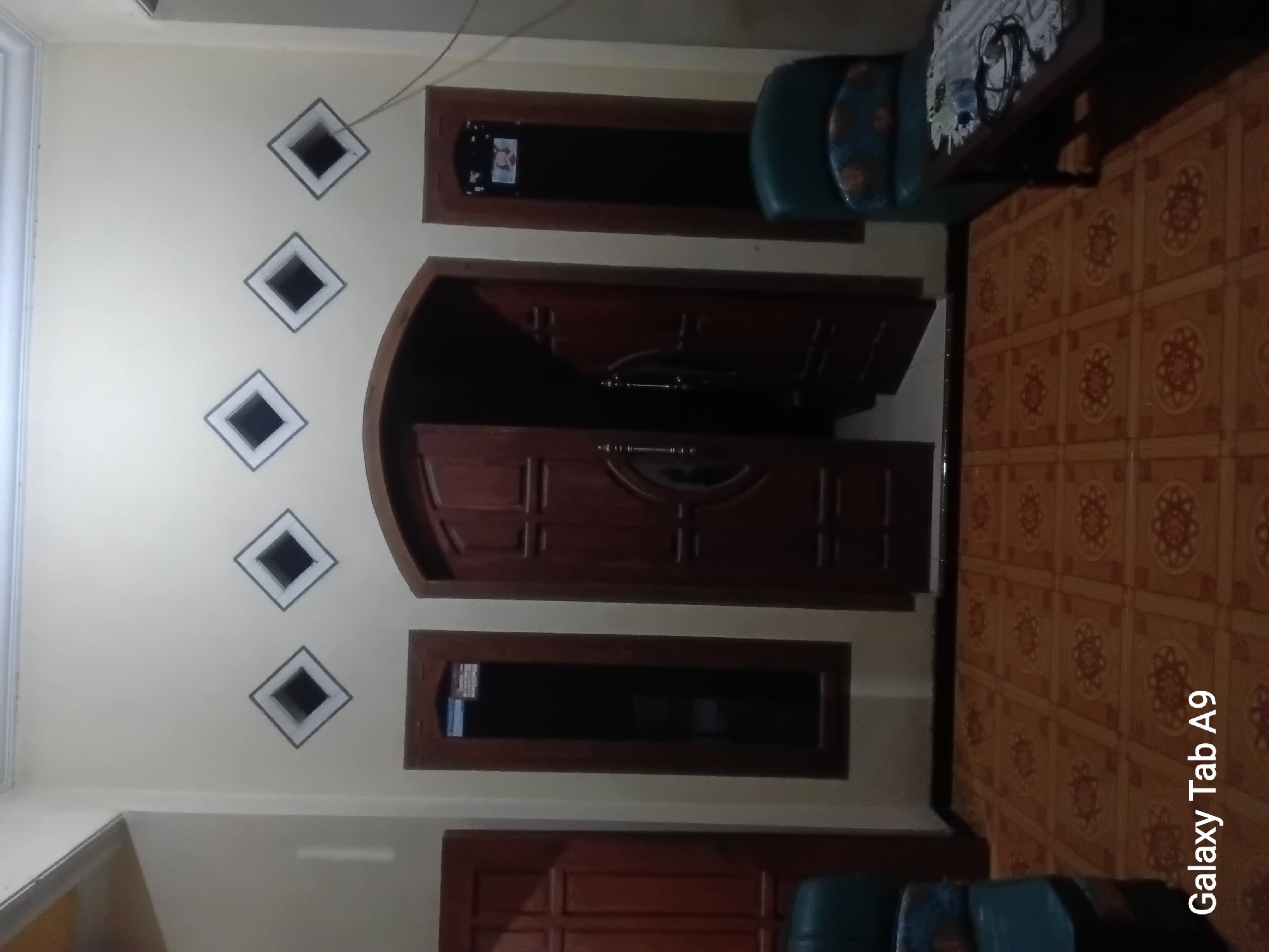 The image depicts an interior view of a room, likely a living room or entryway, with a focus on a large double door in the center. The room has light-colored walls and a patterned tiled floor in shades of brown and orange. Above the central door, there are three diamond-shaped decorative vents. To the left of the double door is a tall, narrow window with a dark pane and a wooden frame. To the right of the double door is another similar tall, narrow window. On the far left of the image, a portion of another wooden door is visible. In the foreground, to the right, is a dark wooden table with a patterned cloth on top, and a dark green armchair.
The image contains the text "Galaxy Tab A9" in the lower left corner, indicating it was taken with a Samsung Galaxy Tab A9 device. There are no people visible in the image. The room appears to be dimly lit, possibly at dusk or night. The overall impression is of a residential interior in Indonesia, possibly Trenggalek, Indonesia, given the stylistic elements and the device used for capture.